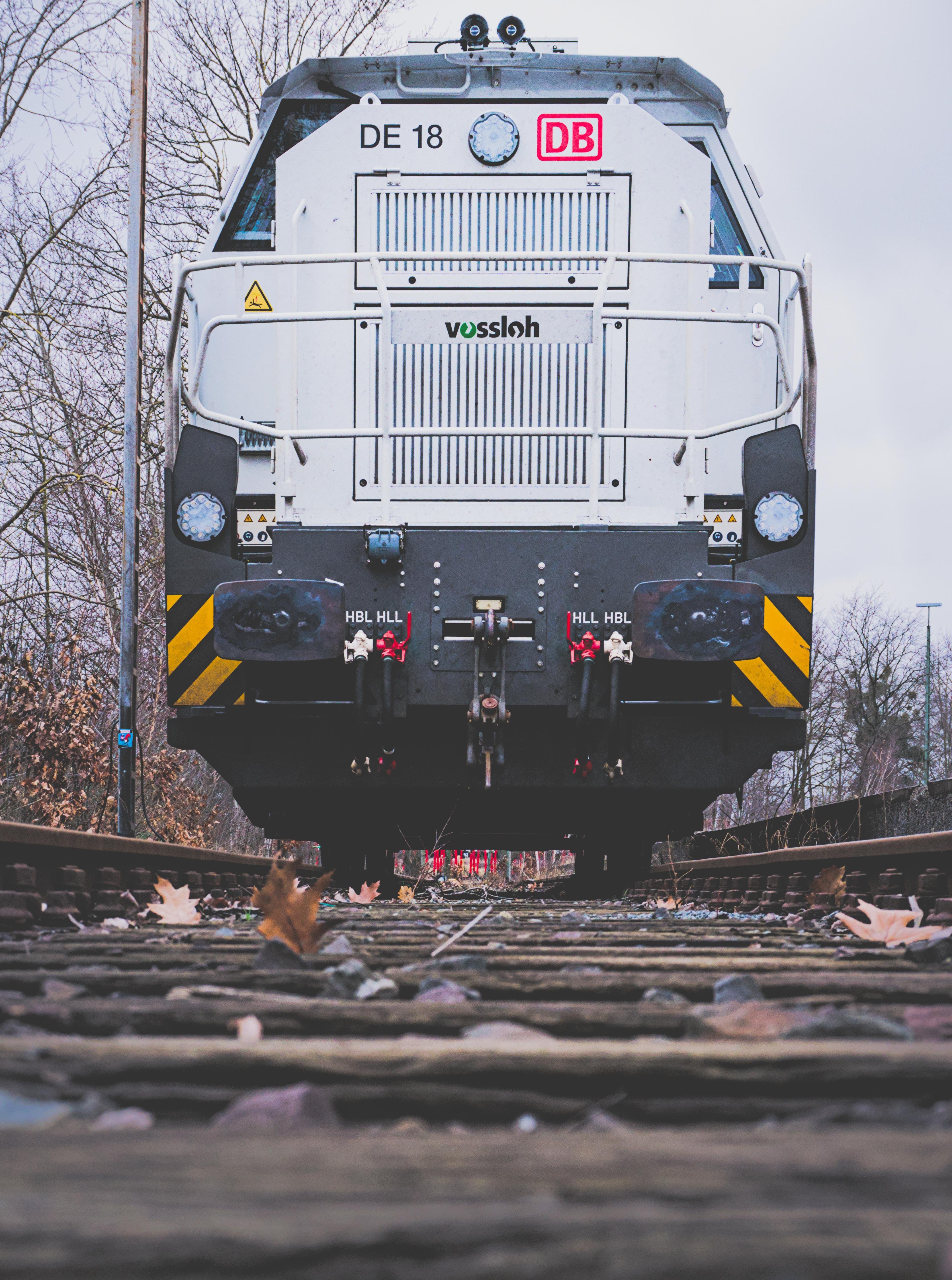 The image shows the front view of a train locomotive on railroad tracks. The locomotive is marked with "DE 18" and "DB," with the brand "Vossloh" visible. The perspective is low, close to the ground, with the tracks and some fallen leaves in the foreground. Trees are visible in the background.
