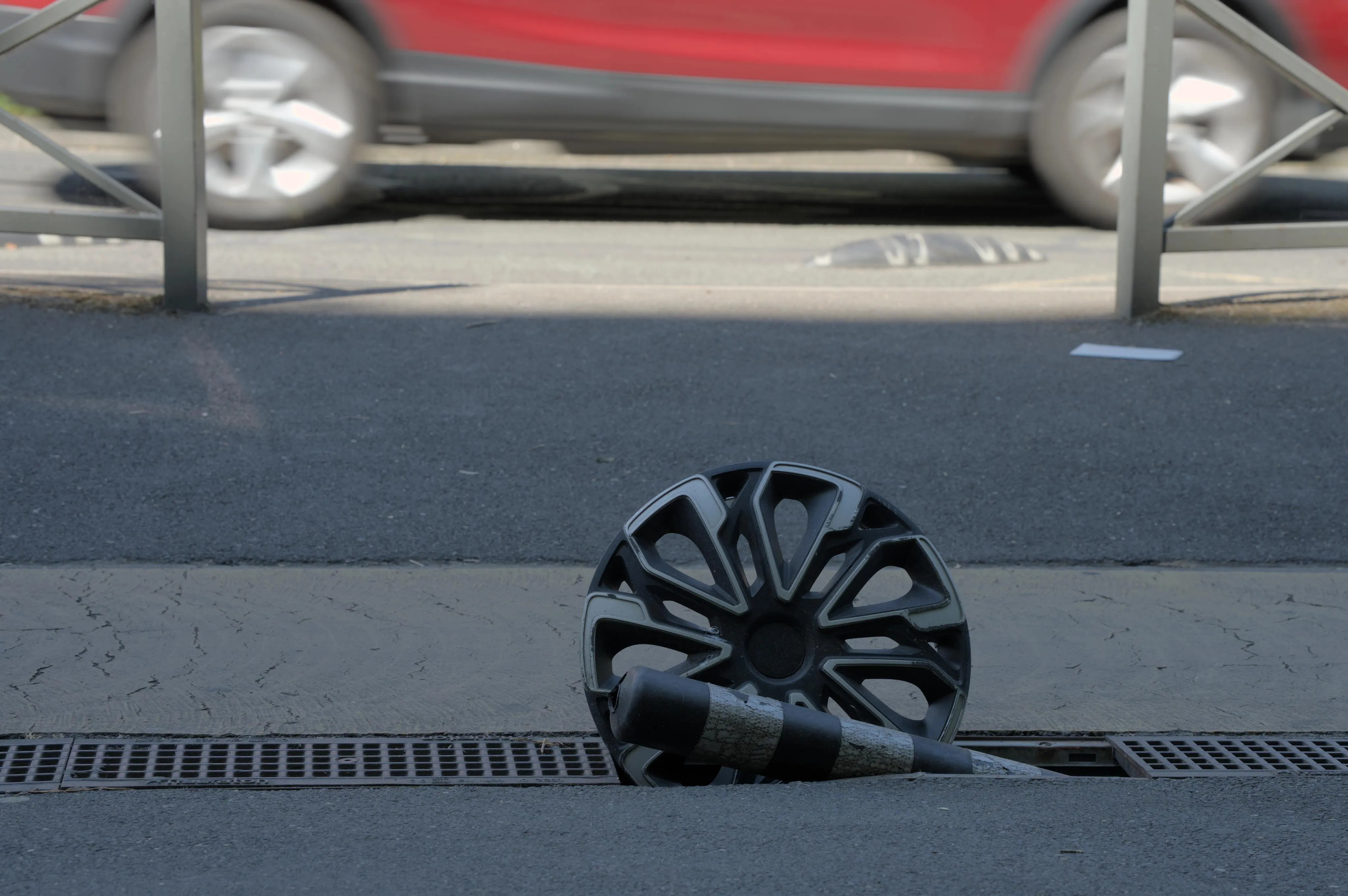 Photograph of an automobile wheel rim planted in the middle of the drain in a sidewalk, along with a broken cylindrical roadblock plot. The wheel rim has traces of wear and tear. The sidewalk is in the shade. In the background on the sunlit road, the wheels and bottom part of the red body of a moving car are blurred by the car's motion.