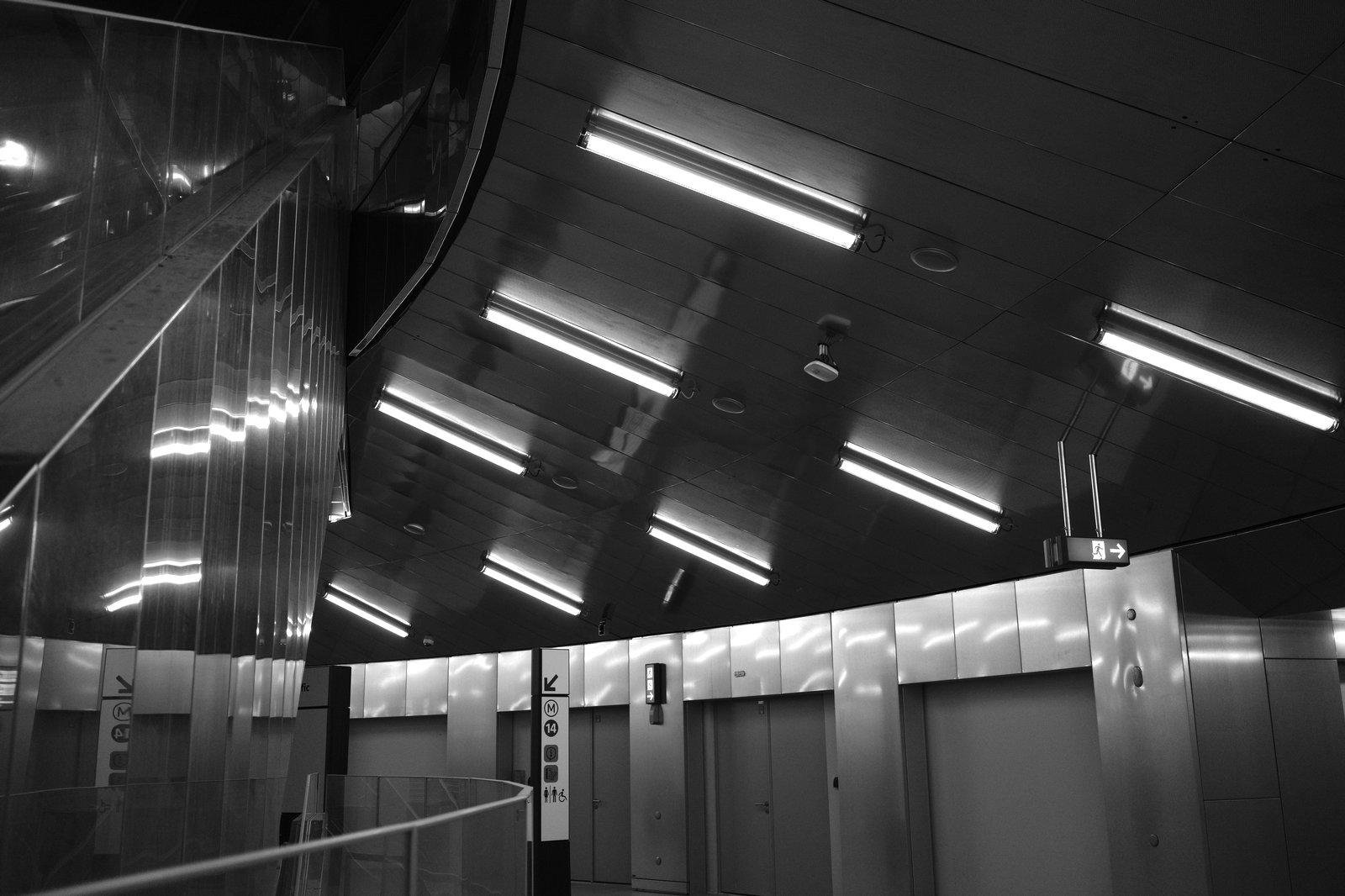 Photo horizontale noir et blanc. Un large passage tournant dans une gallerie dont de multiple emplacements pour magasin ont leur rideau de fer baissés, éclairé par des barres de LED comme des tubes néons. Une paroie en métal réfléchissant d’un escalier est à gauche