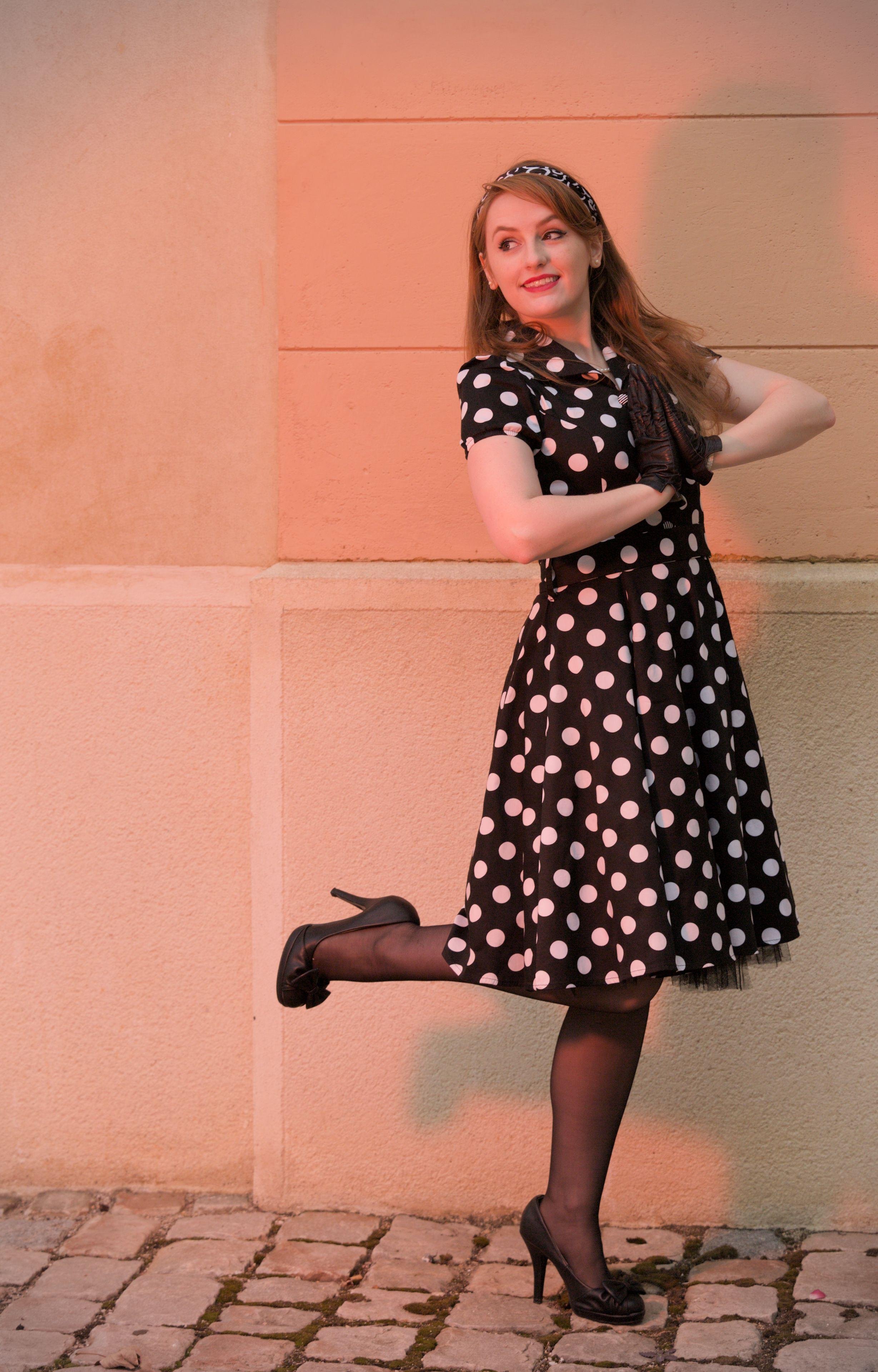 Photo format portrait montrant une jeune femme rousse aux cheveux long, coiffée d’un bandeau à la mode année 50, noir et blanc, une robe noire à gros pois blanc, des chaussures noires à talon haut, se tenant sur sa jambe gauche, pliant la jambe droite pour former un angle à 90⁰ avec sa cuisse. Les mains gantées de noir sont jointes à plat comme pour une prière, elle regarde vers l’arrière avec un sourire de joie. La scène est dans les teintes rouge/orange