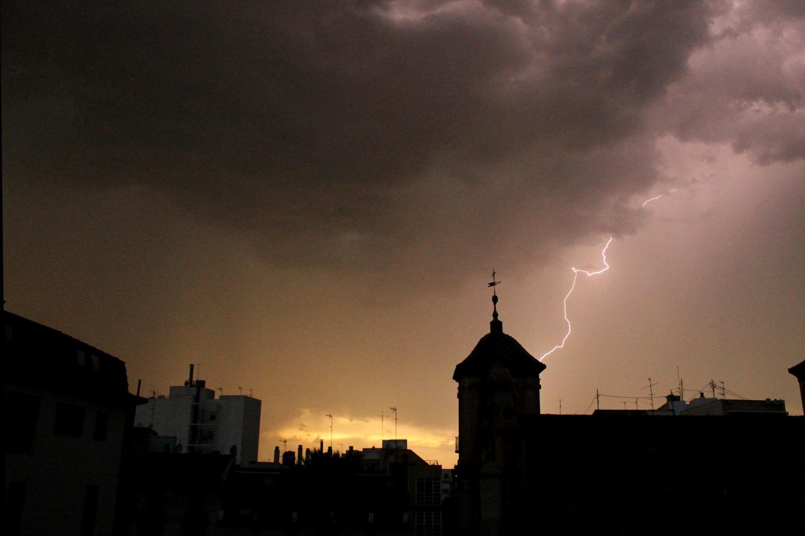Foto de un paisaje urbano donde los edificios se ven a contra luz y se aprecia una torre-campanario de una iglesia, con un cielo de puesta de sol nublado por nubes muy oscuras de tormenta. En el cielo anaranjado se ve una parte de las nubes iluminadas por un rayo que está cayendo justo sobre la silueta de la torre de la iglesia
