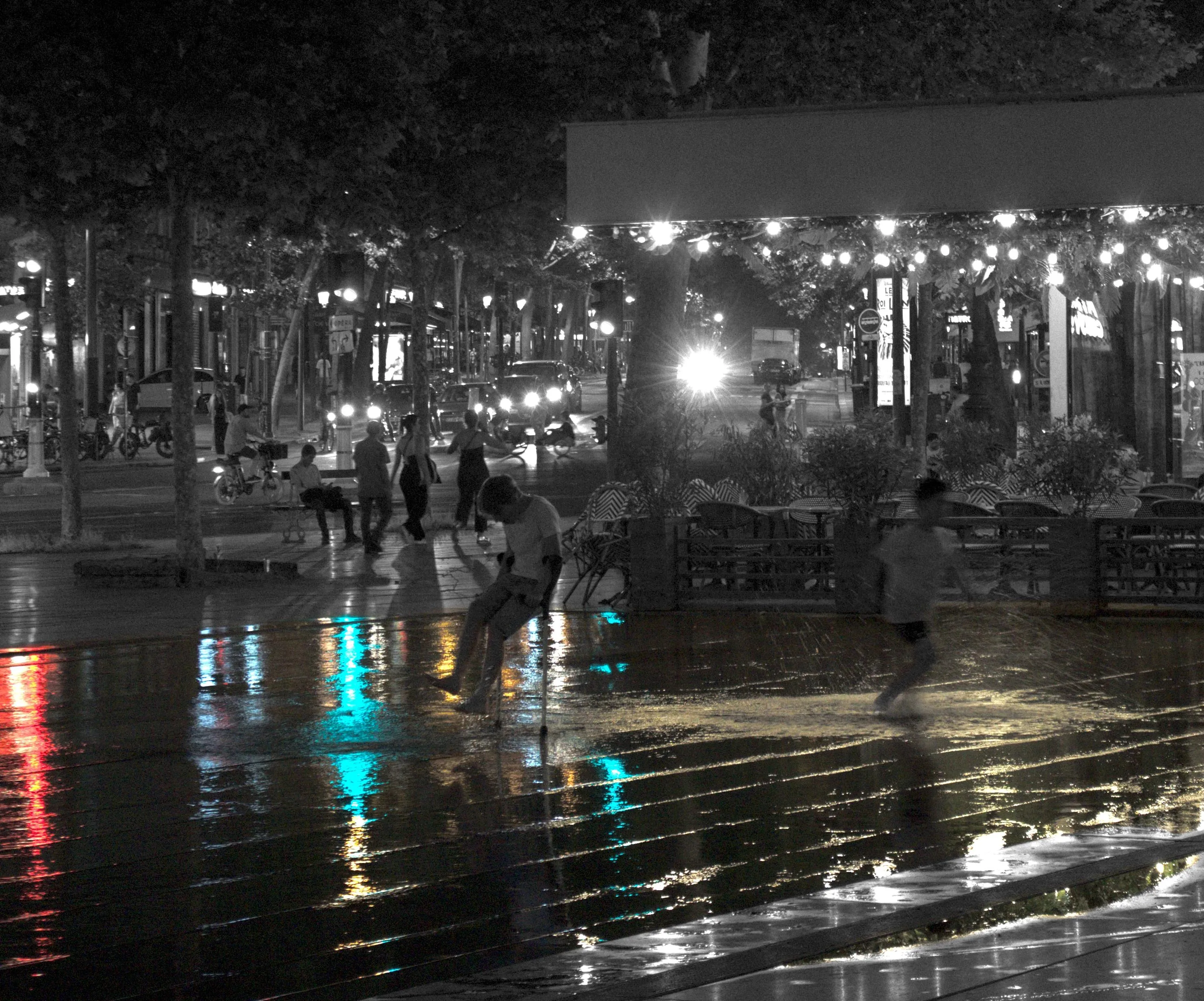 Photograph of children playing around on a public square. Most of the surface of the square visible in the image is covered in water. City lights are reflected on the water surface. One of the kids is using crutches and made a water splash. The other kid seems to be running away from the first. The surface of the water has a green coloration, reflecting the trees surrounding the square. The reflected city lights form bright red, yellow and blue columns on the water surface. The rest of the photograph is in monochrome, including the children that are detoured.