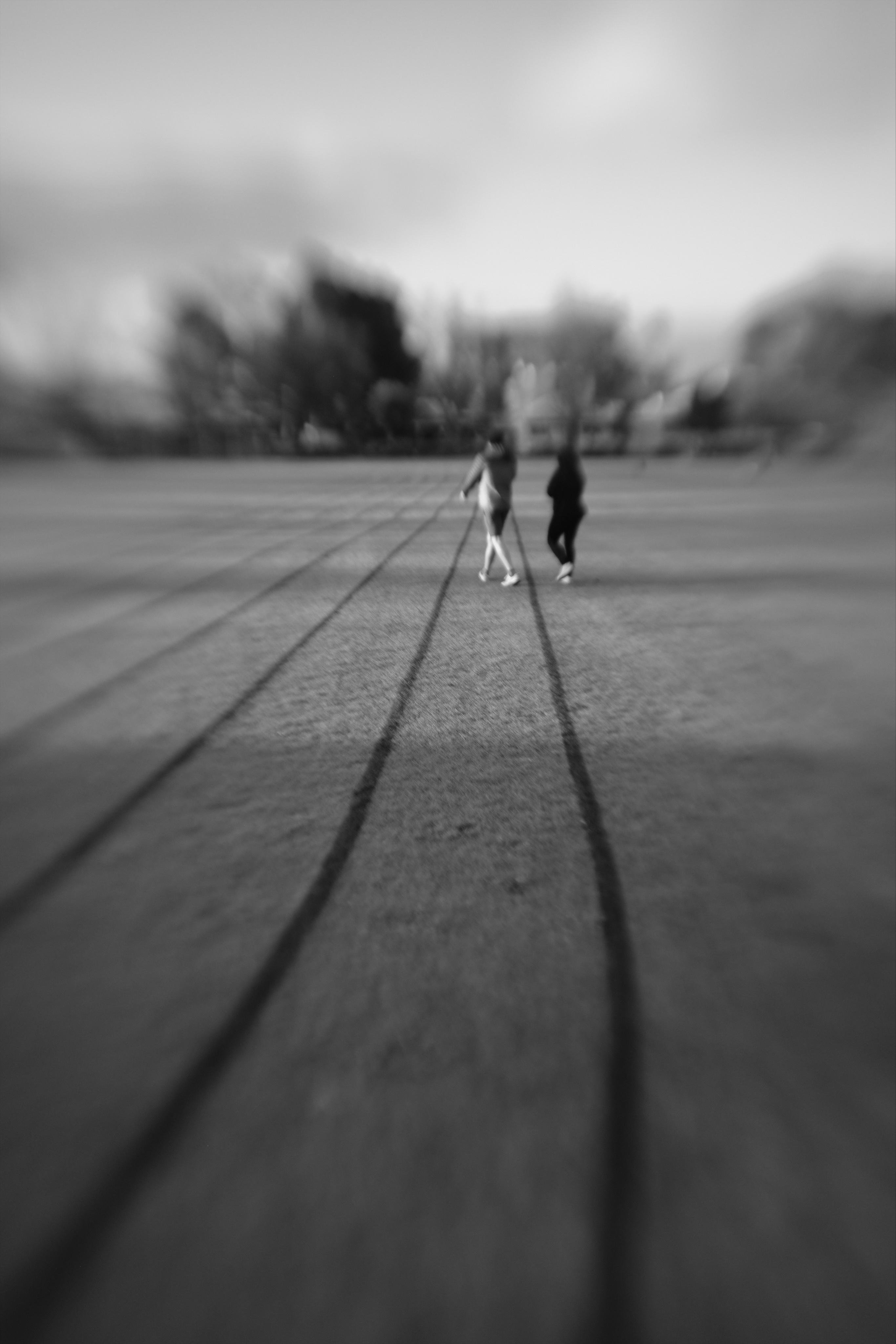 Black-and-white photo of two people walking away on a wide open field with long, parallel shadows stretching toward the camera; trees and sky in the distance are heavily blurred.