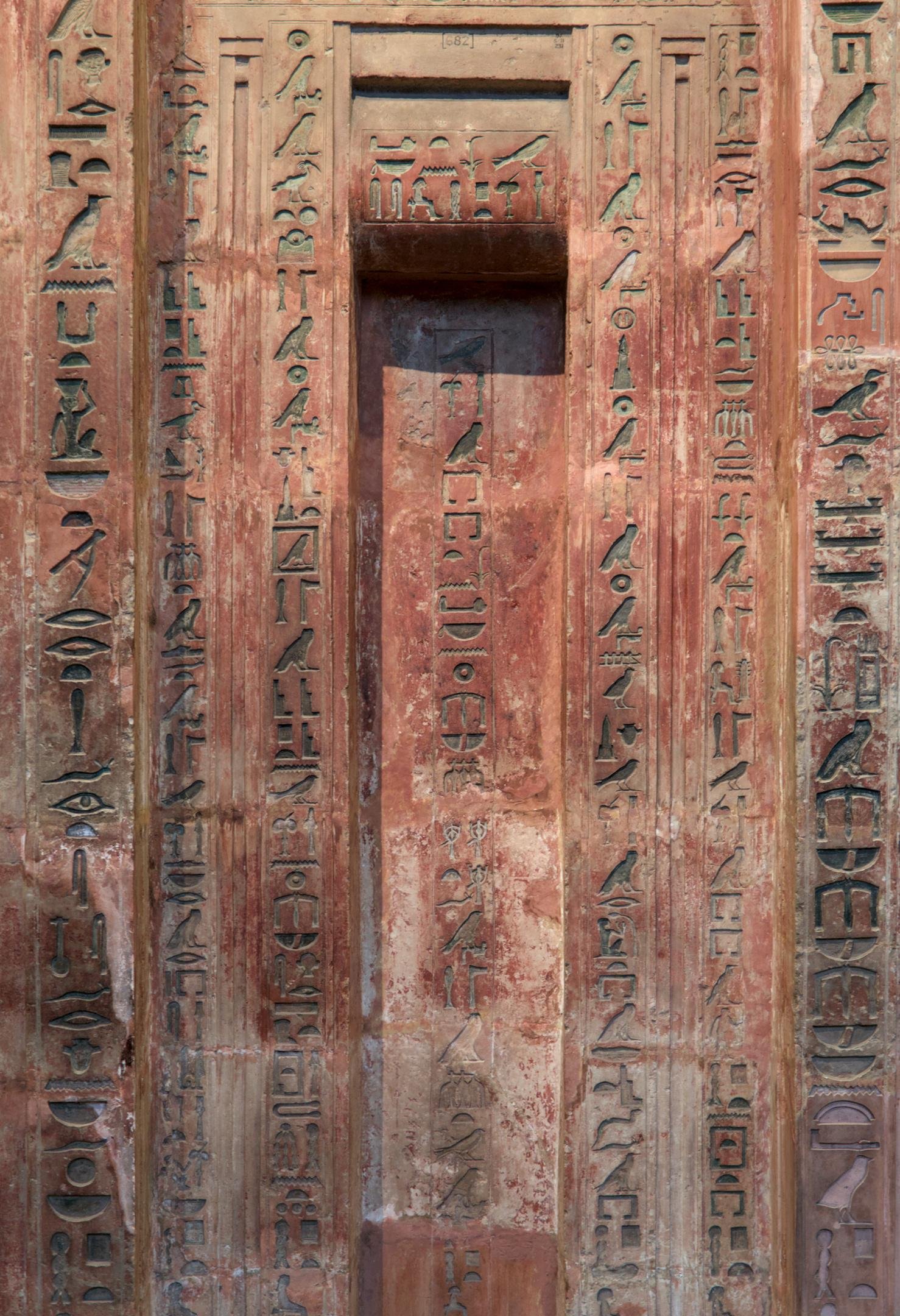 Photo verticale. Vue de face d’une porte symbolique avec plusieurs cartouches verticaux plein de hieroglyphes. L’ensemble forme des arche rectangulaire concentrique en allant vers le centre, donnant du relief et de couleur ocre