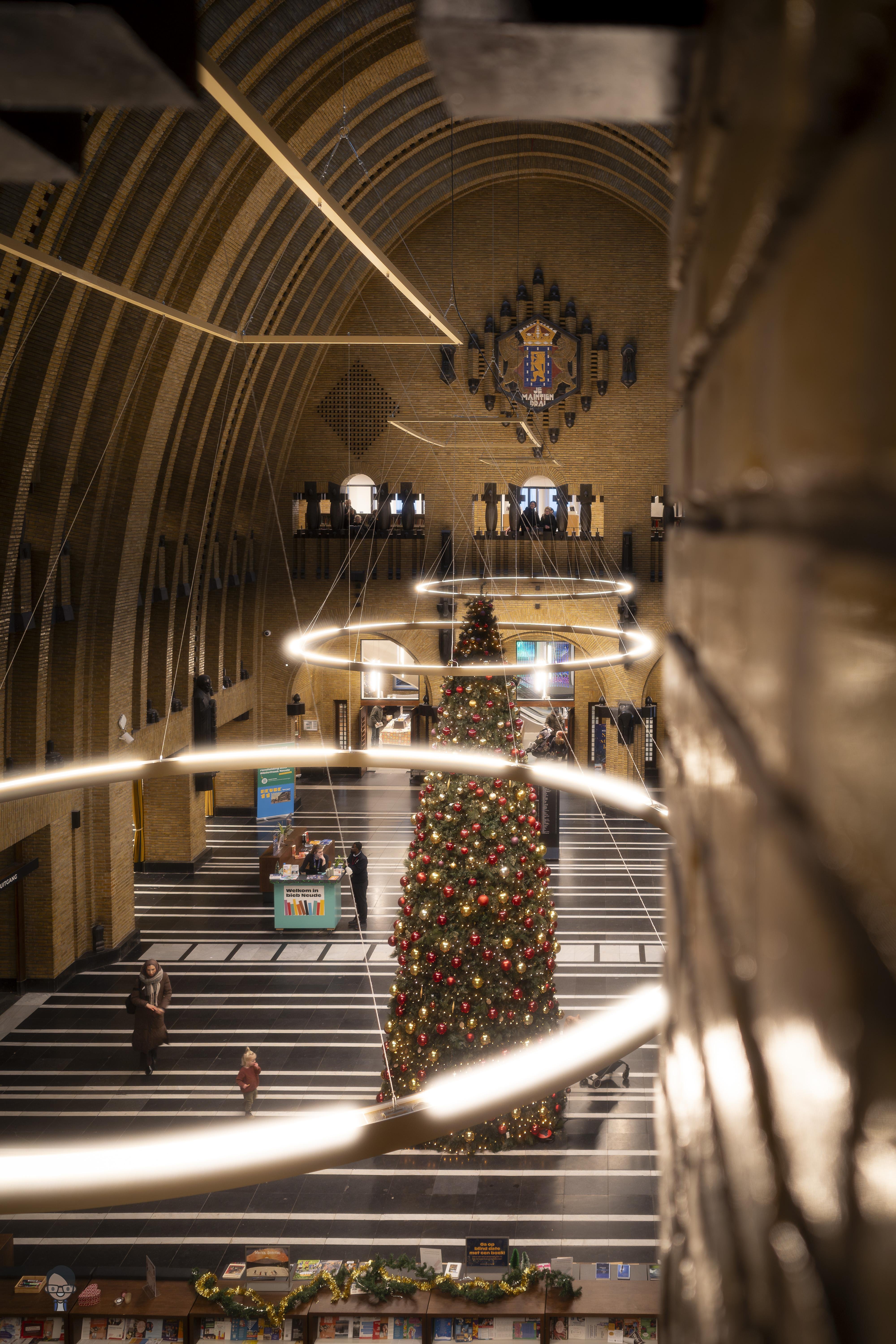 The image shows the interior of a large, elegantly designed building with high, arched ceilings. There is a decorated Christmas tree with lights and ornaments in the center of a spacious hall. Suspended circular lights add to the festive atmosphere. People can be seen walking around the area. The building has intricate architectural details and a crest or emblem on the wall above.