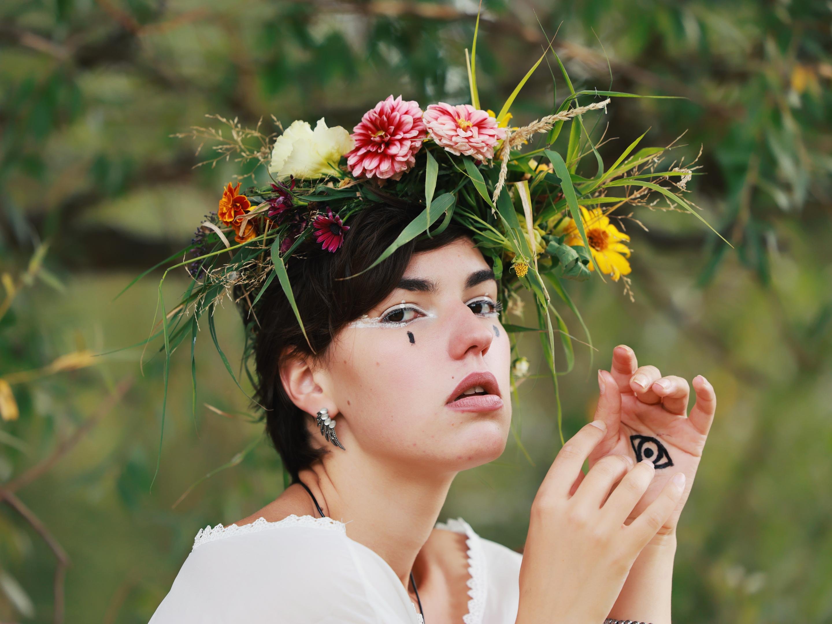 Person wearing a flower crown made of various colorful flowers, such as daisies and chrysanthemums, with green foliage, set against a blurred natural background. Facial features include artistic makeup with drawn black tear-shaped elements near the eyes and white eyeliner. A small eye symbol is painted on the back of the right hand. The person wears a white garment and has short dark hair with silver earrings.