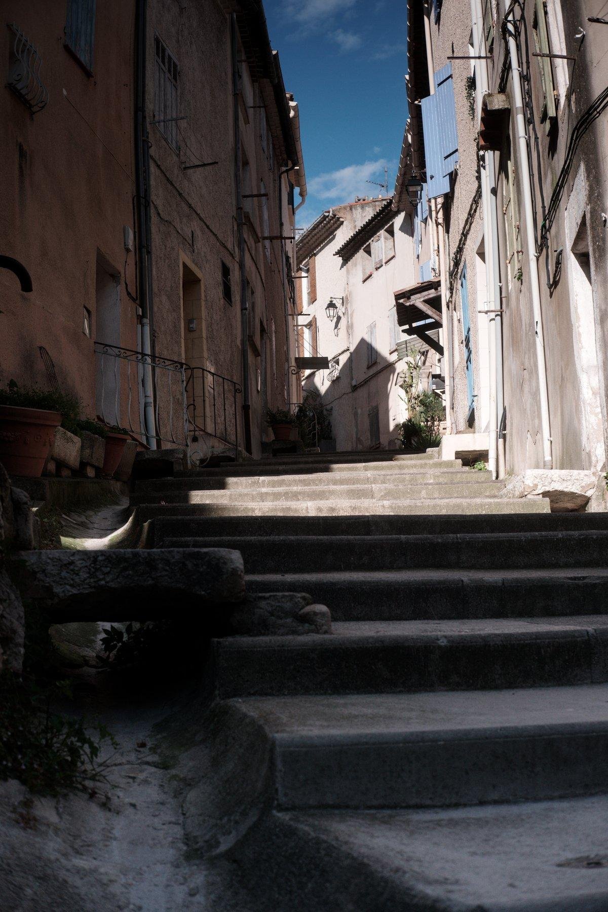 Photo verticale. Un chemin piéton qui monte entre des maison, commençant par quelques marches. Sur la gauche, un canal d’évacuation d’eau. Entre les maisons, on voit le ciel bleu