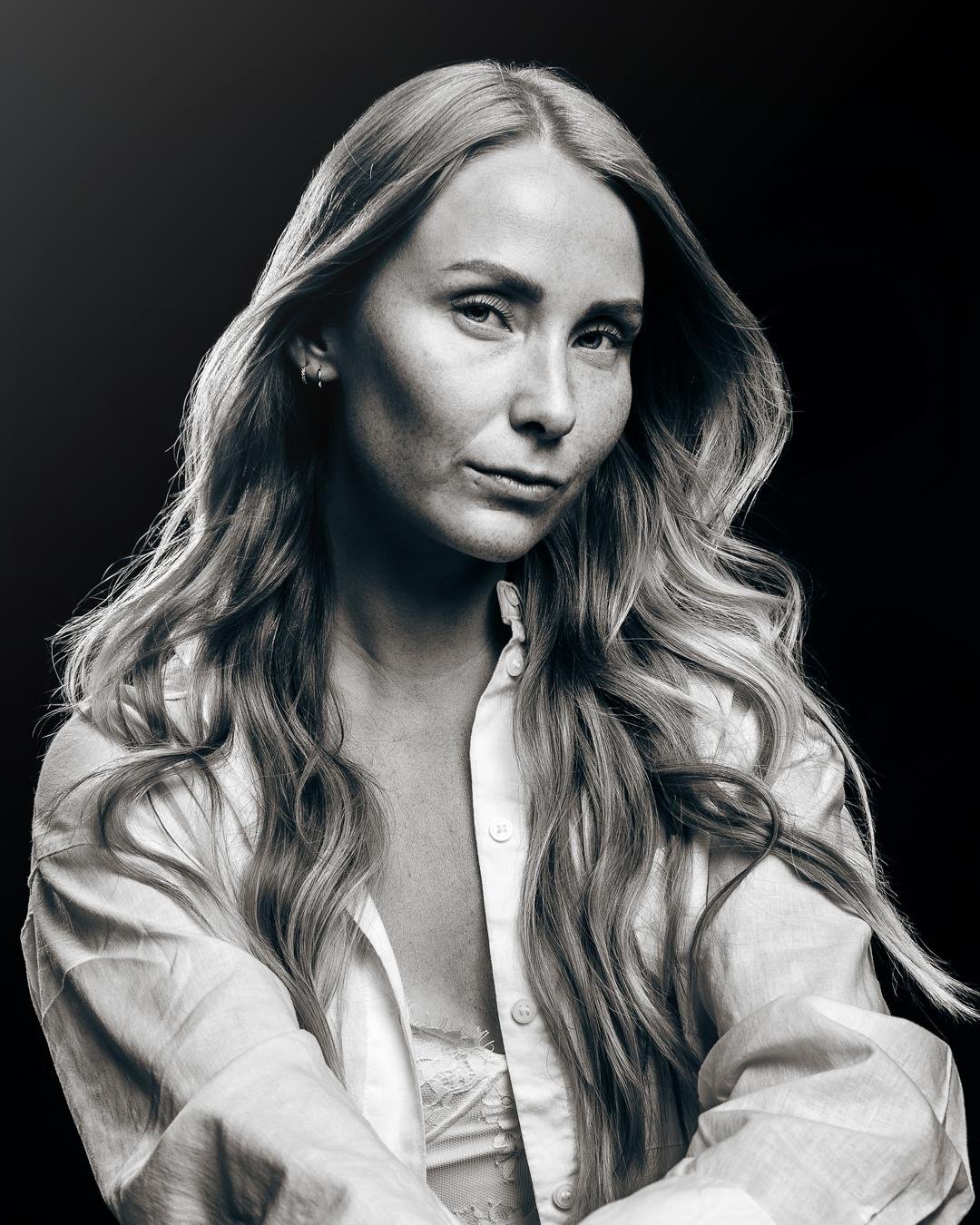 A black and white fine art portrait of a woman with long, wavy hair. She gazes confidently at the camera, wearing a white button-down shirt over a lace top. The soft lighting enhances the contours of her face and the delicate details of her clothing, creating a timeless and elegant composition.