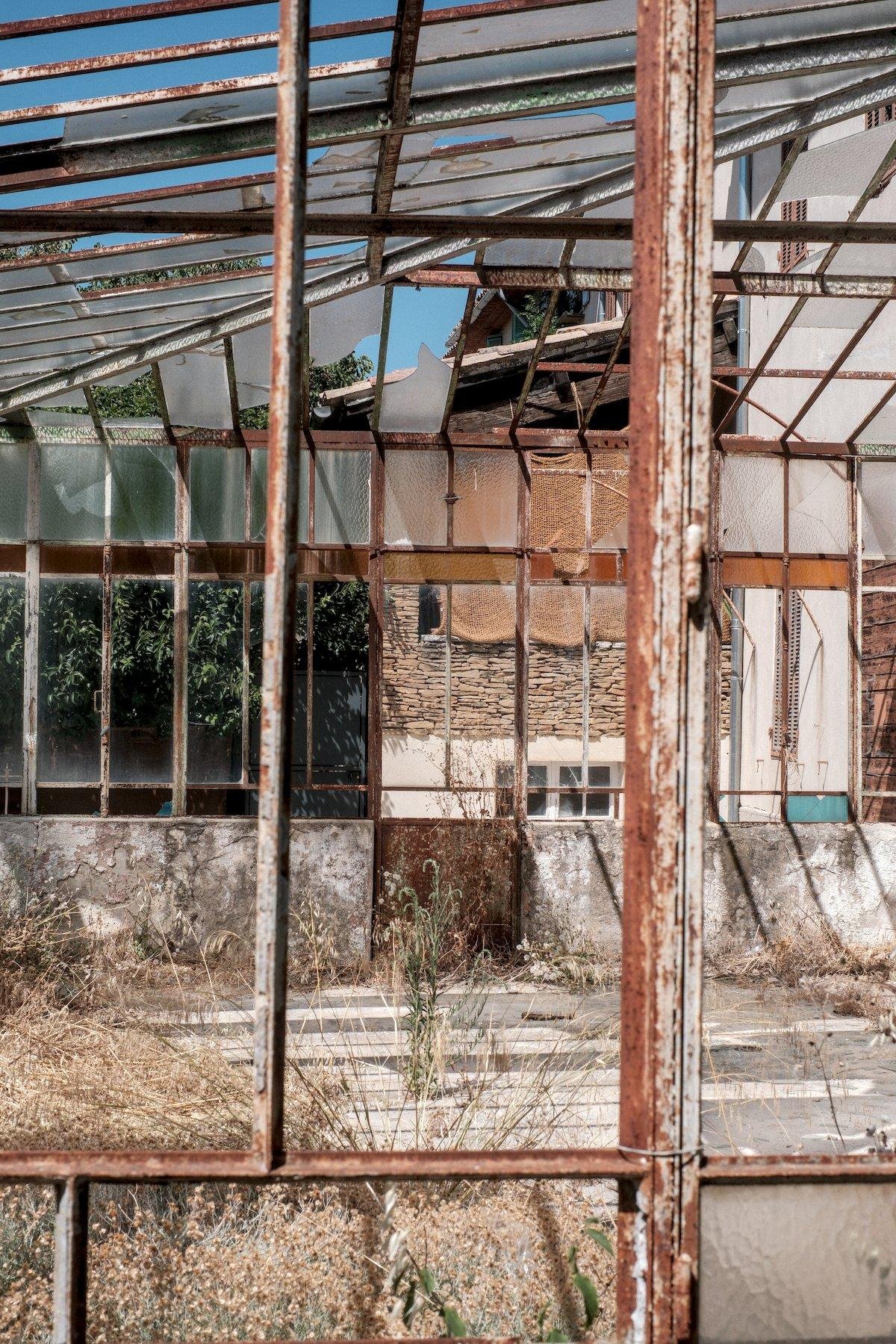 Photo verticale. À travers l’armature rouillée d’une veranda à l’abandon, à la verrière brisée, on voit une porte rouillée sur le mur opposé, sans vitre
