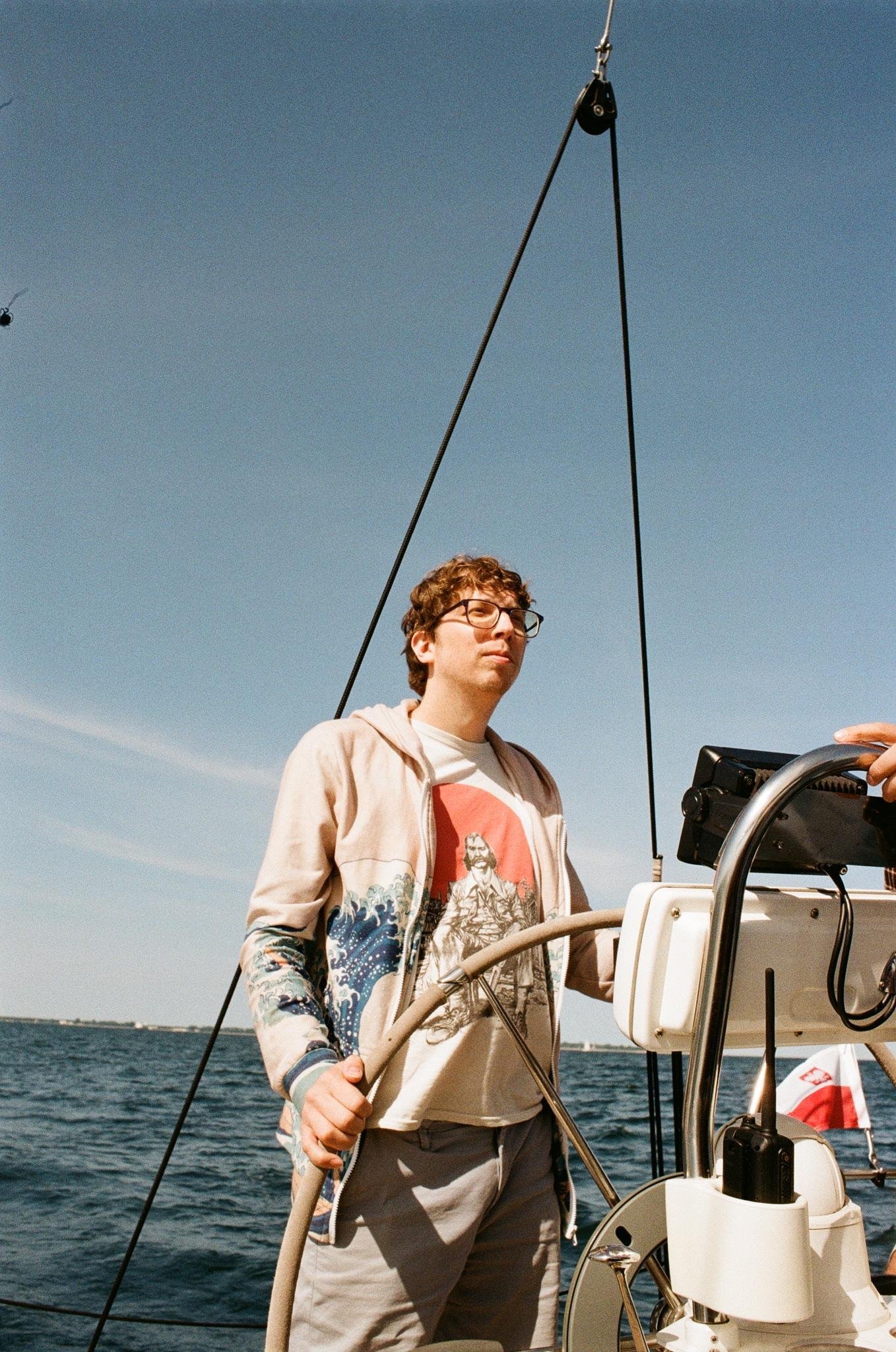 Man wearing glasses stands at the helm of a sailboat, holding the steering wheel with open water and a clear blue sky behind; rigging lines and navigation equipment frame the right side.