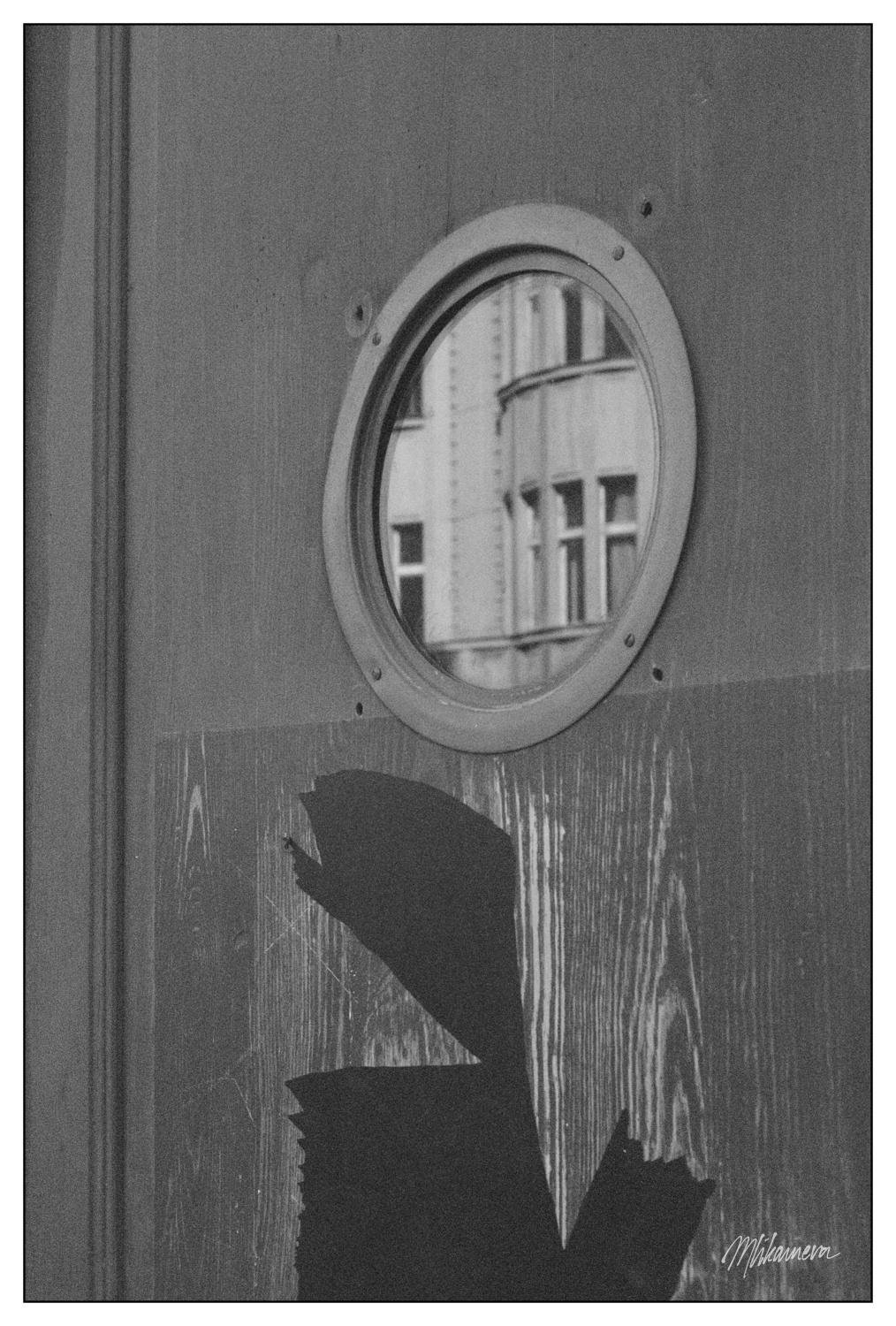 A monochrome photograph of a small round port-like window in a modern wooden door with remains of hastily removed wrap; there is a reflection of a building in said window, the reflection is a series of a small windows on a prewar building