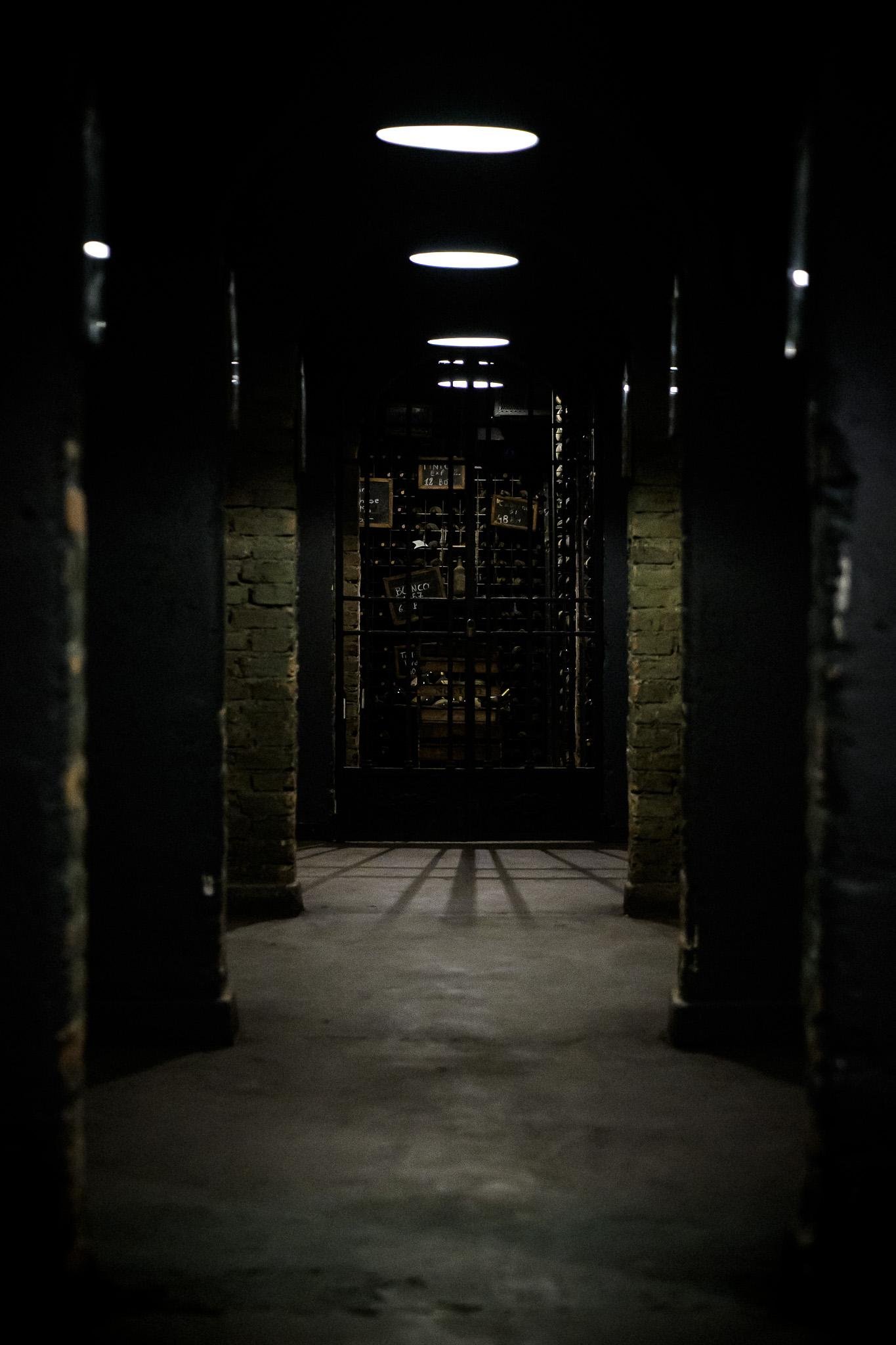 The image depicts a dimly lit hallway leading to a barred gate in a cellar. The lighting above creates a dramatic and somewhat eerie atmosphere. It looks like a secure storage area for wine.