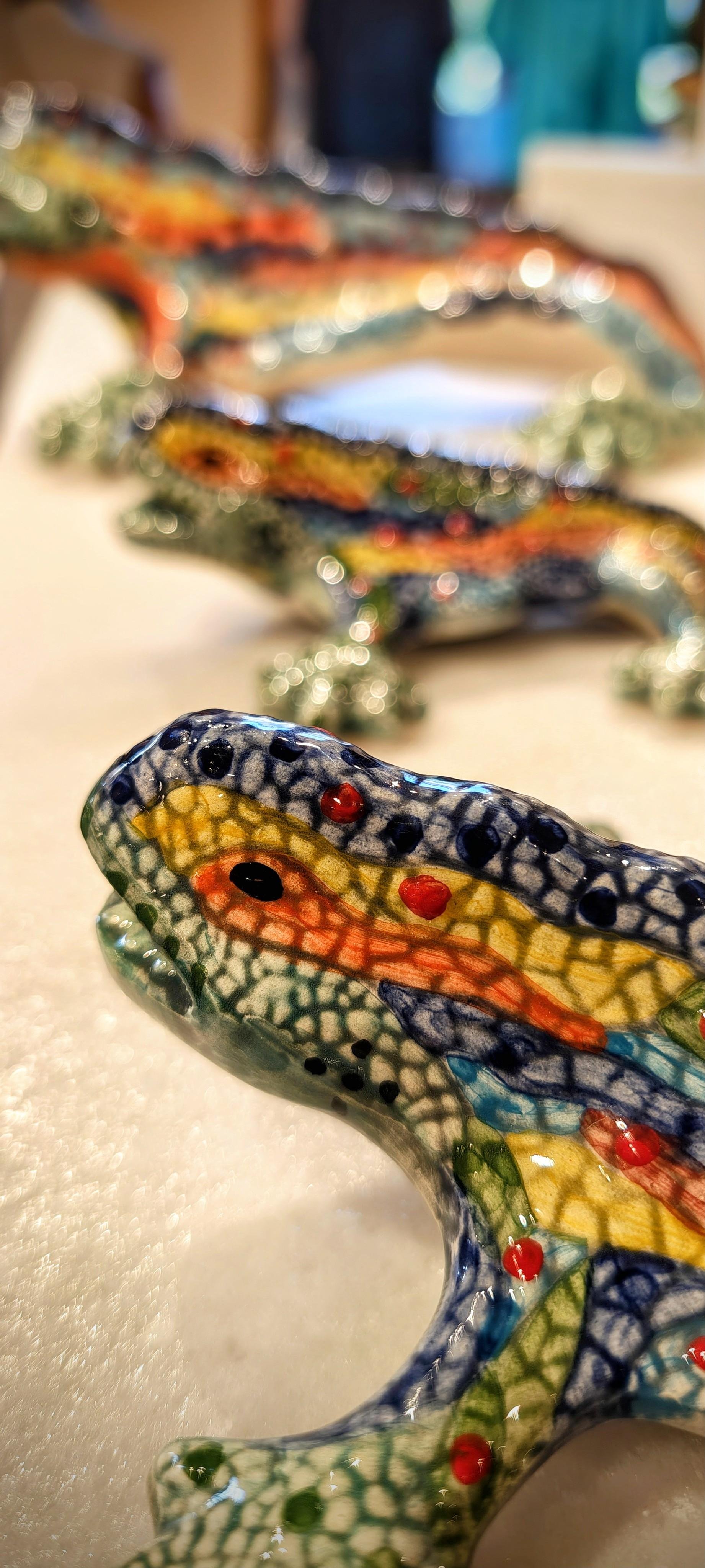 Serie di lucertole in ceramica smaltata dai colori vivaci (blu, giallo, verde, rosso), disposte in fila su un tavolo, con dettagli in primo piano e sfondo sfocato in un ambiente interno
