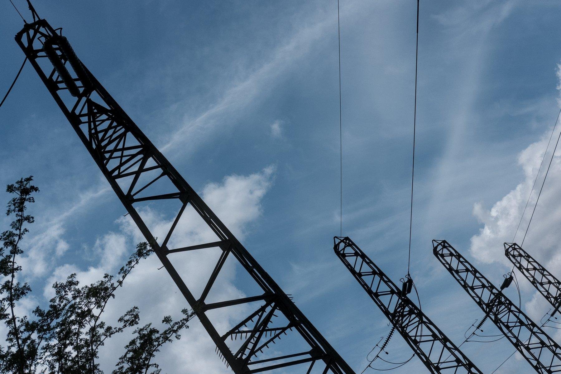 Photo horizontale. Contre-plongée penchée sur des pylônes électriques de haute tension, sous un ciel bleu avec des volutes de nuages