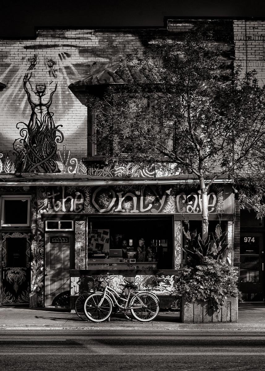 Black-and-white street view of a brick storefront covered in murals and graffiti, with the sign “the only cafe” above an open window counter; a bicycle is parked on the sidewalk in front, and a leafy tree and planter partially block the right side of the facade.