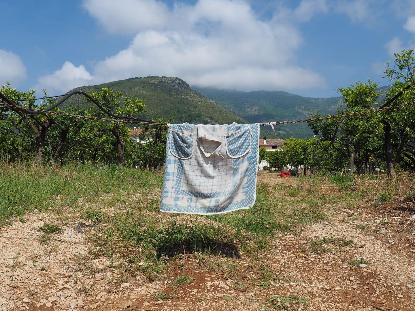 Un vecchio grembiule, lacero, steso su una catena posta all'ingresso di un terreno privato, davanti a una casetta di campagna. Il grembiule non sembra avere altro scopo che rendere più visibile la catena. In sfondo, delle montagne sovrastate da un cielo sereno, seppur solcato da nuvole poco preoccupanti.