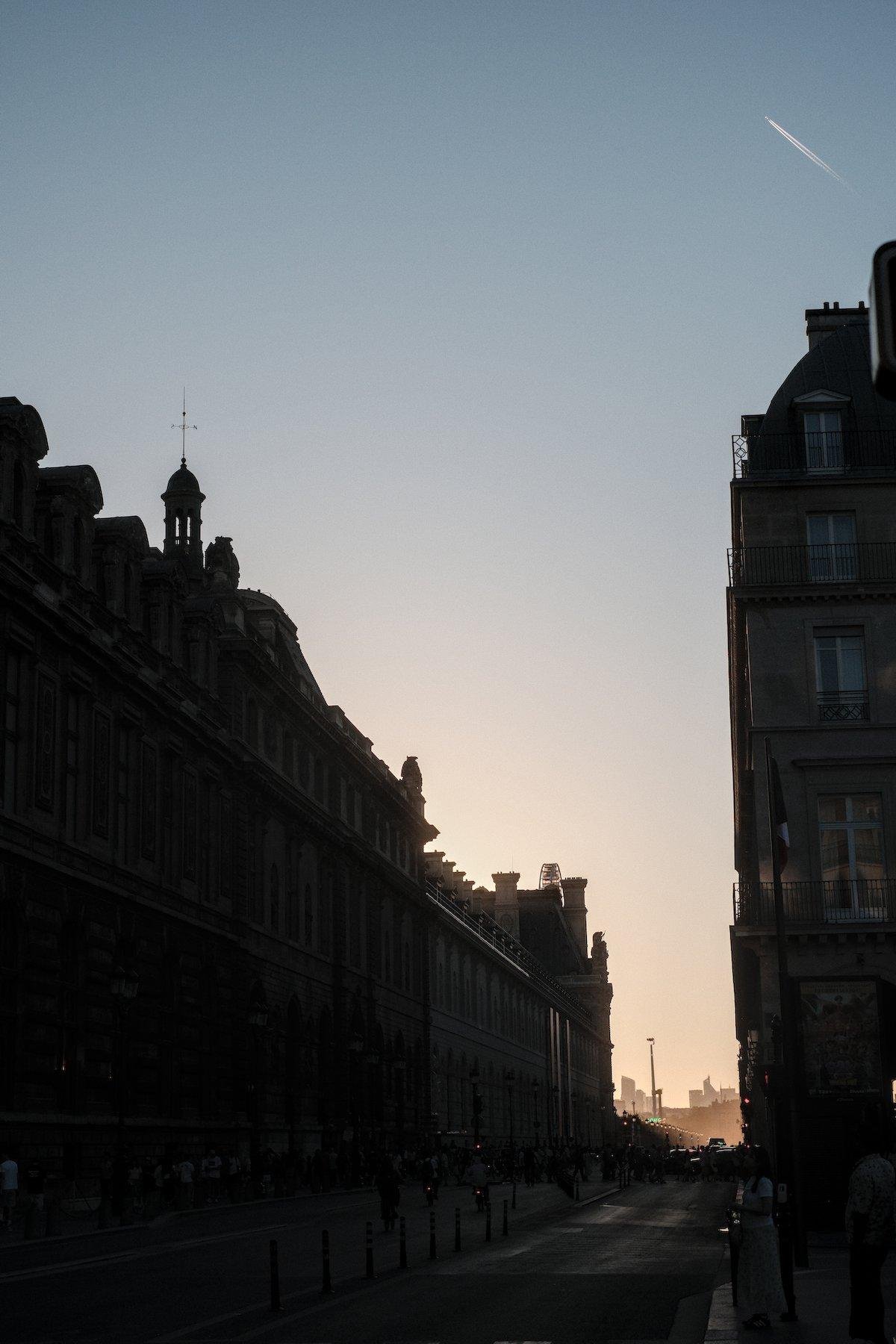 Photo verticale. Une rue parisienne bordée d’immeuble de 4 ou 5 étages, en contre jour, sous un ciel de soleil couchant dérangé par la trace d’un avion en train de passer