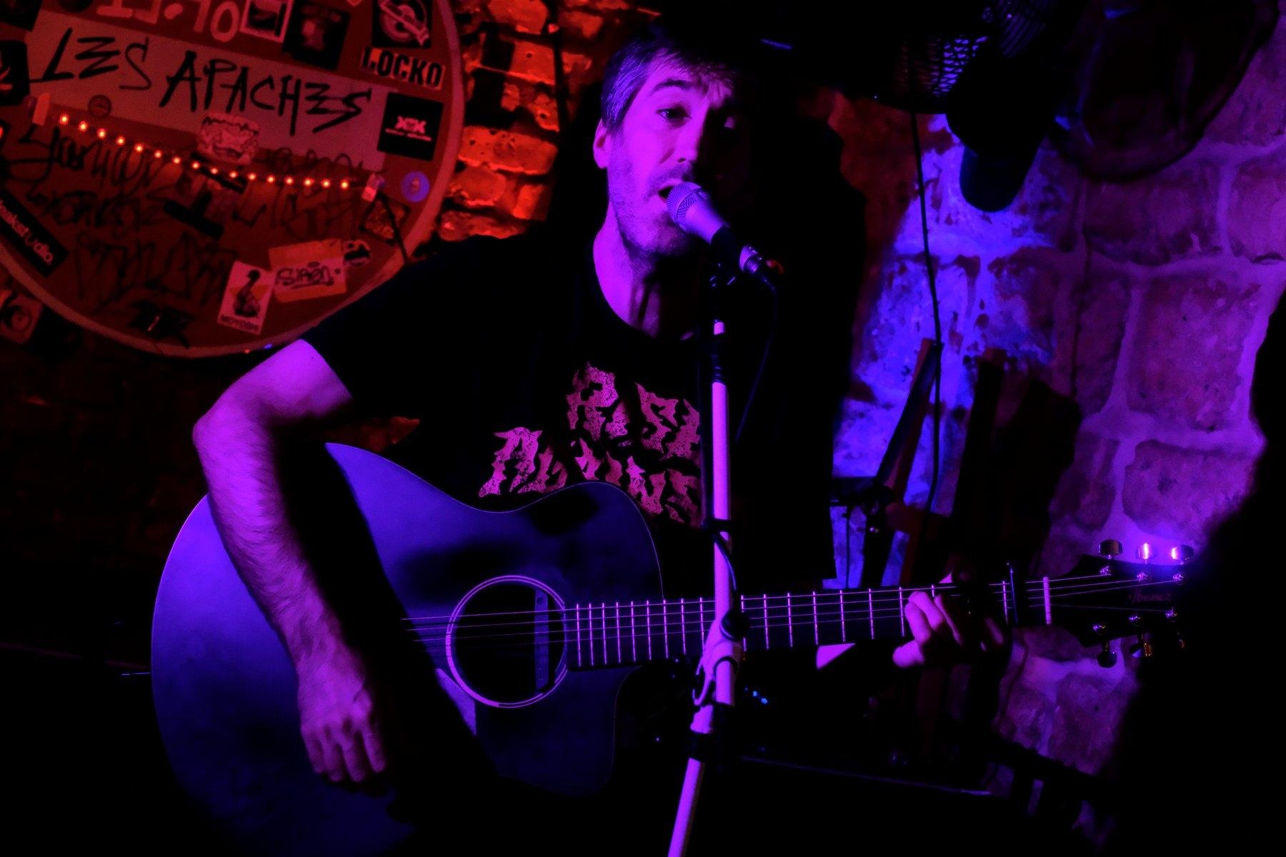 Photo horizontale. Un homme jouant de la guitare et chantant dans un micro. Le mur derrière lui est éclairé en rouge, rendant le panneau Stop très sombre, et le mur à droite est éclairé en violet. Le musicien est un mélange de teinte de violet selon où la lumière atterrie