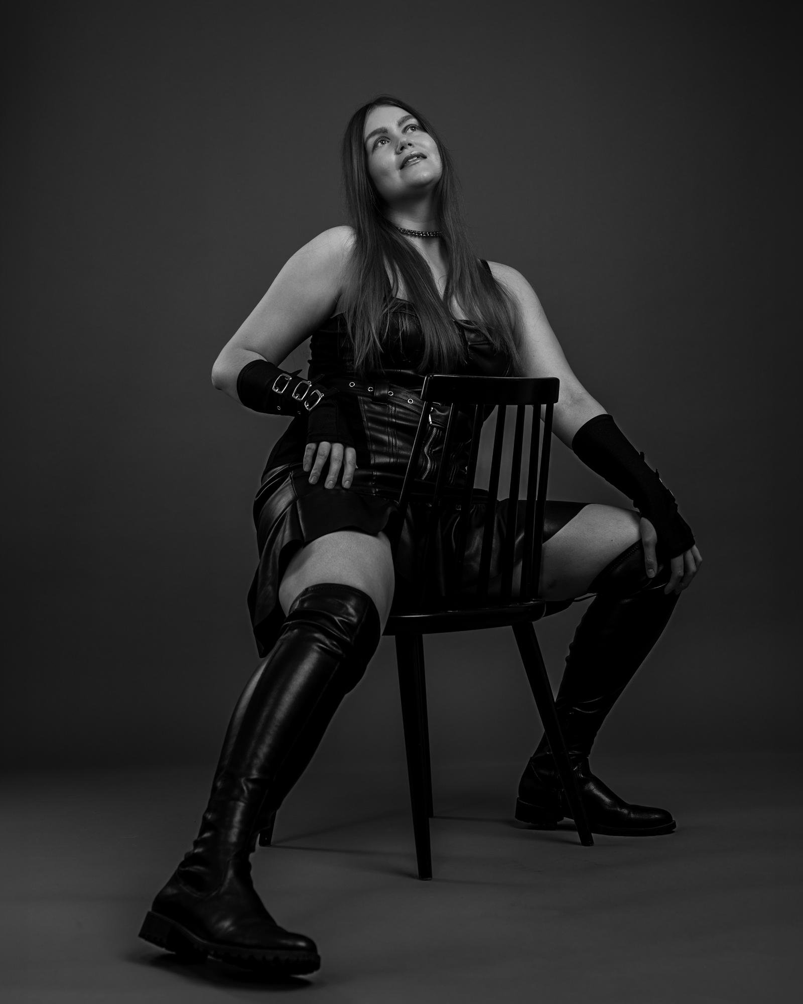 The image is a black and white photograph of a person sitting on a chair. They are wearing boots, a dress, and long gloves. The setting has a dramatic, artistic feel with a dark background, and the person is posing in a confident manner.