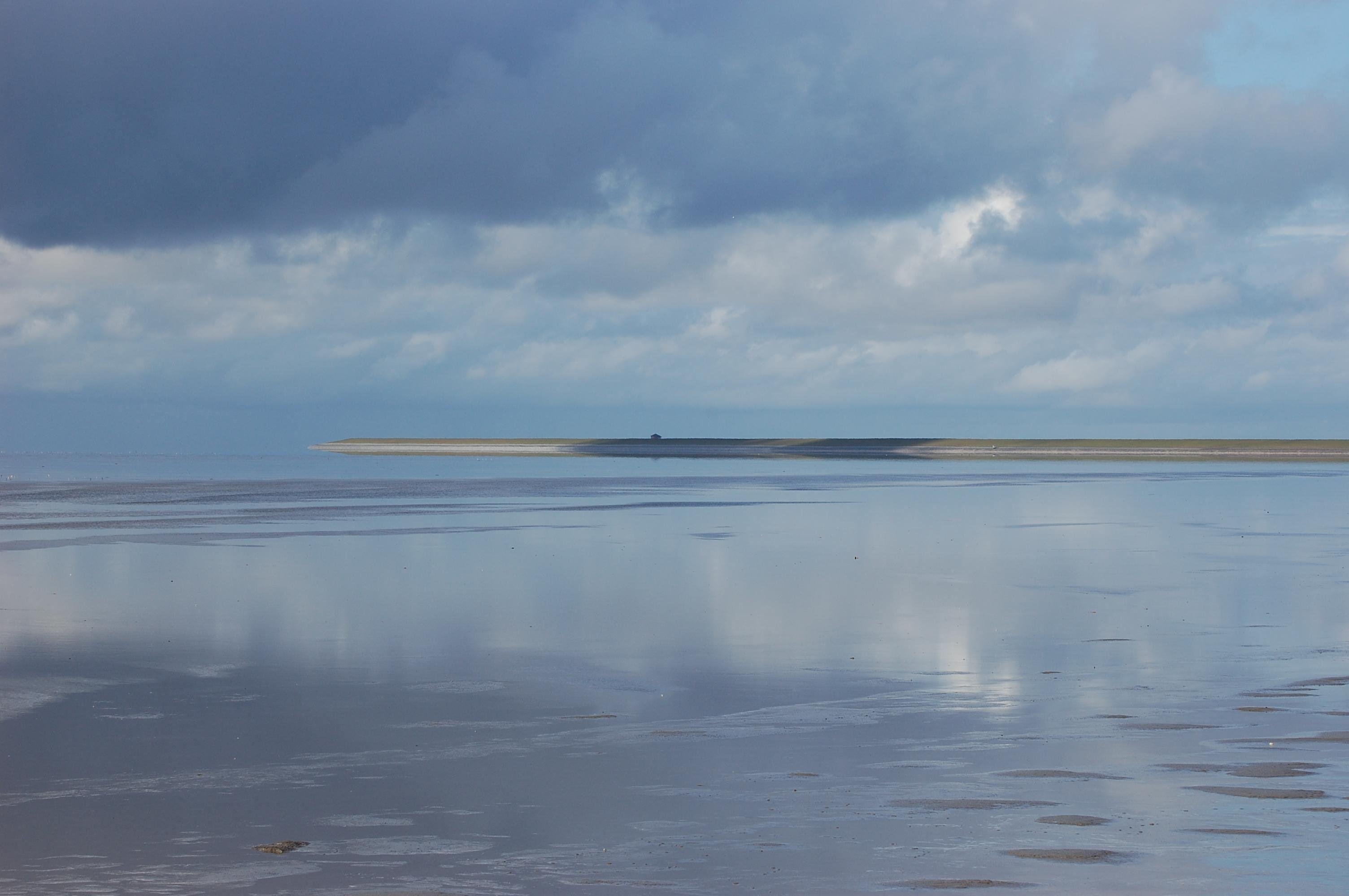 Cloudy sky with dark clouds reflecting on a vast, wet beach. Horizon line shows a thin strip of land in the distance. The surface of the beach is smooth with a few small puddles and ripples.