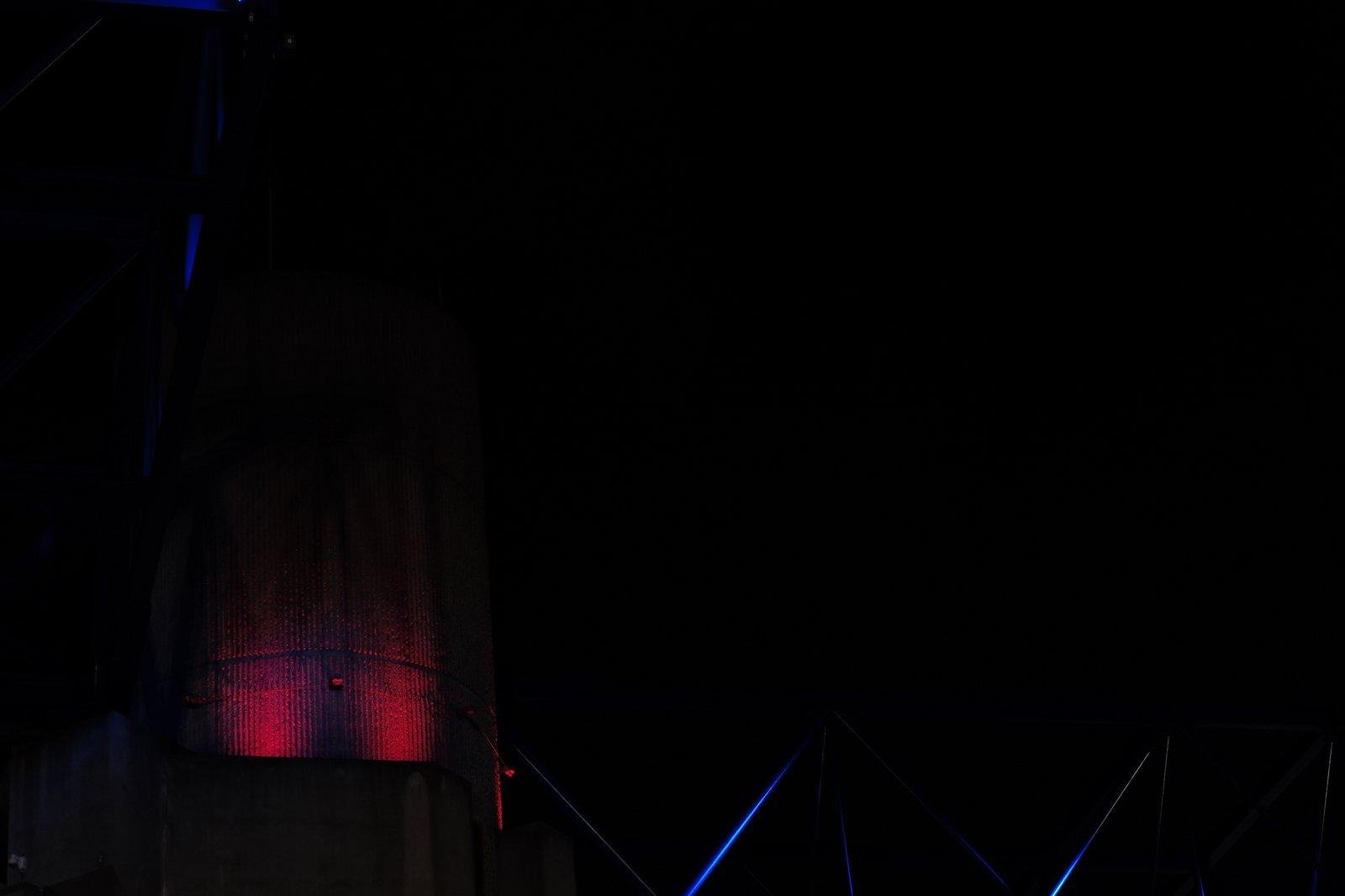 Photo horizontale. Nuit noire, on voit surtout des lampes rouges éclairant un mur, vers le haut, et des traits lumineux bleu