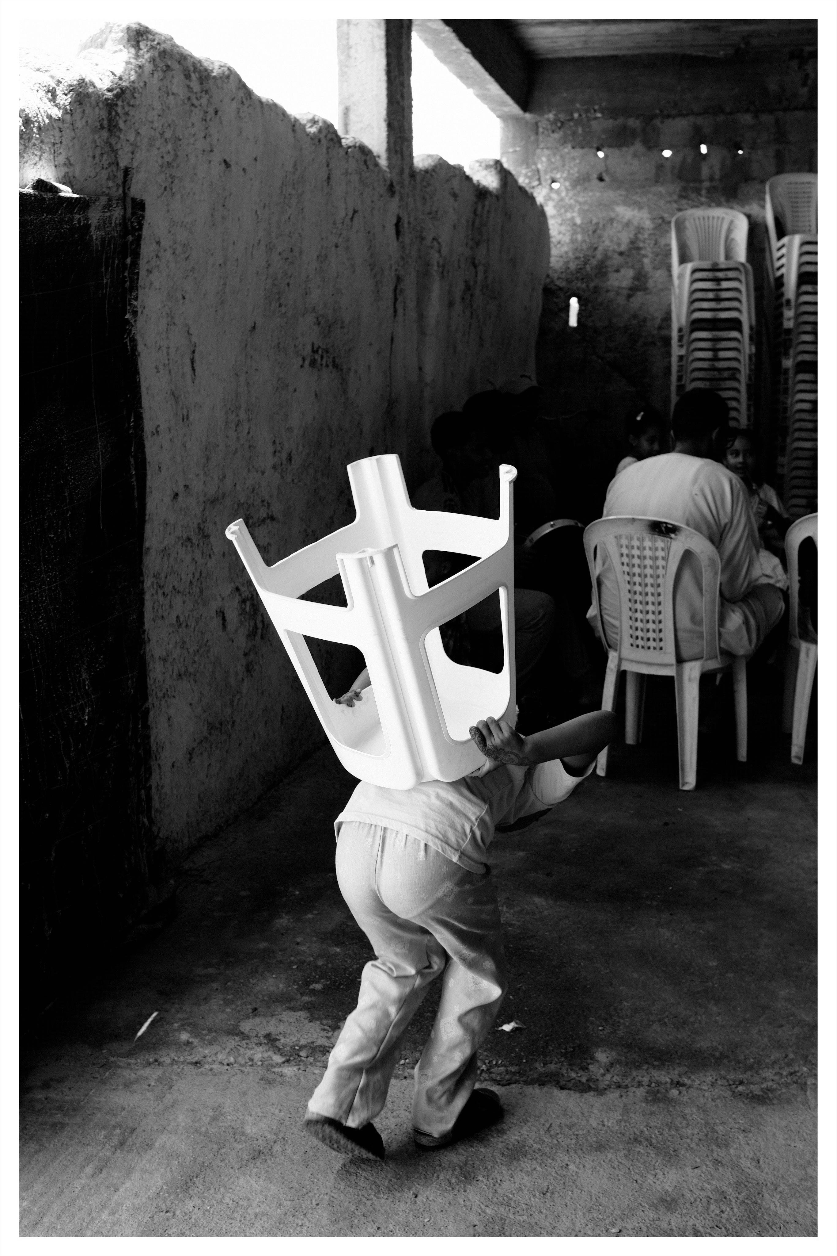 Black-and-white photo of a small child walking away in a narrow concrete courtyard, carrying an upside-down plastic chair over their head; stacked plastic chairs and seated people are in shadow under a roof on the right, with a rough plaster wall along the left.