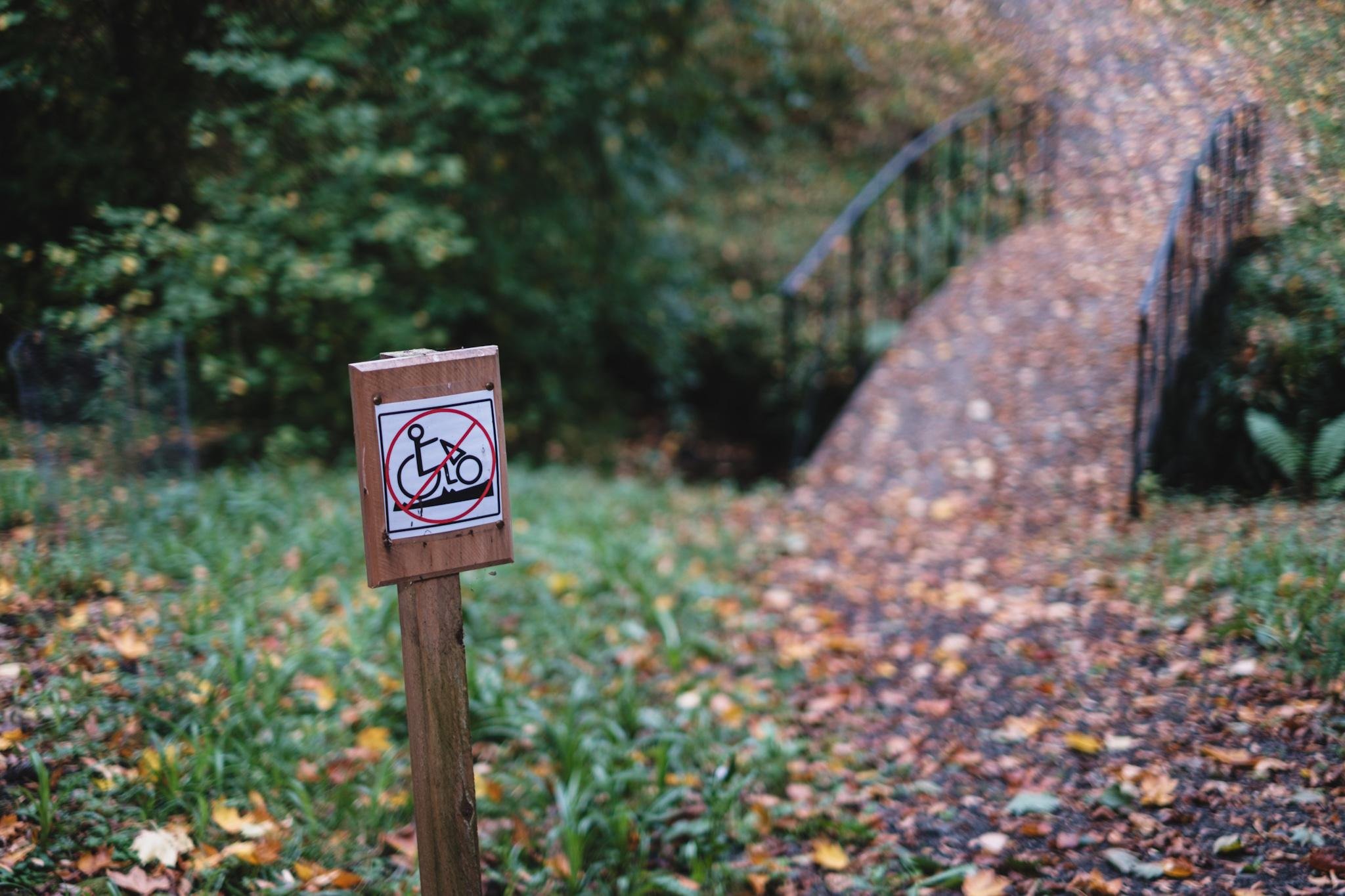 A wooden post with a sign prohibiting mobility scooters, set in a grassy area with fallen leaves. In the background, a curved footbridge covered in autumn leaves crosses over a stream.