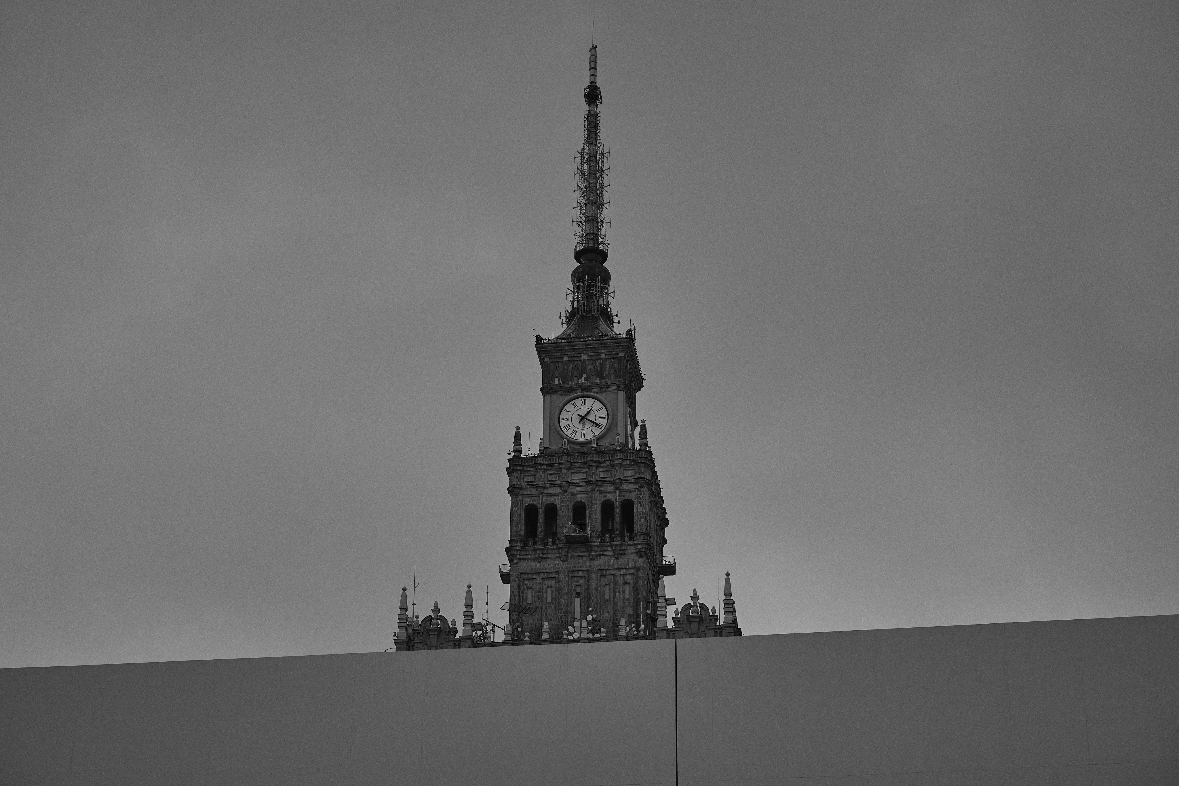 The image features the upper part of the Palace of Culture and Science in Warsaw, Poland. It's a prominent building with a clock and an intricate spire, showcased against a gray sky.