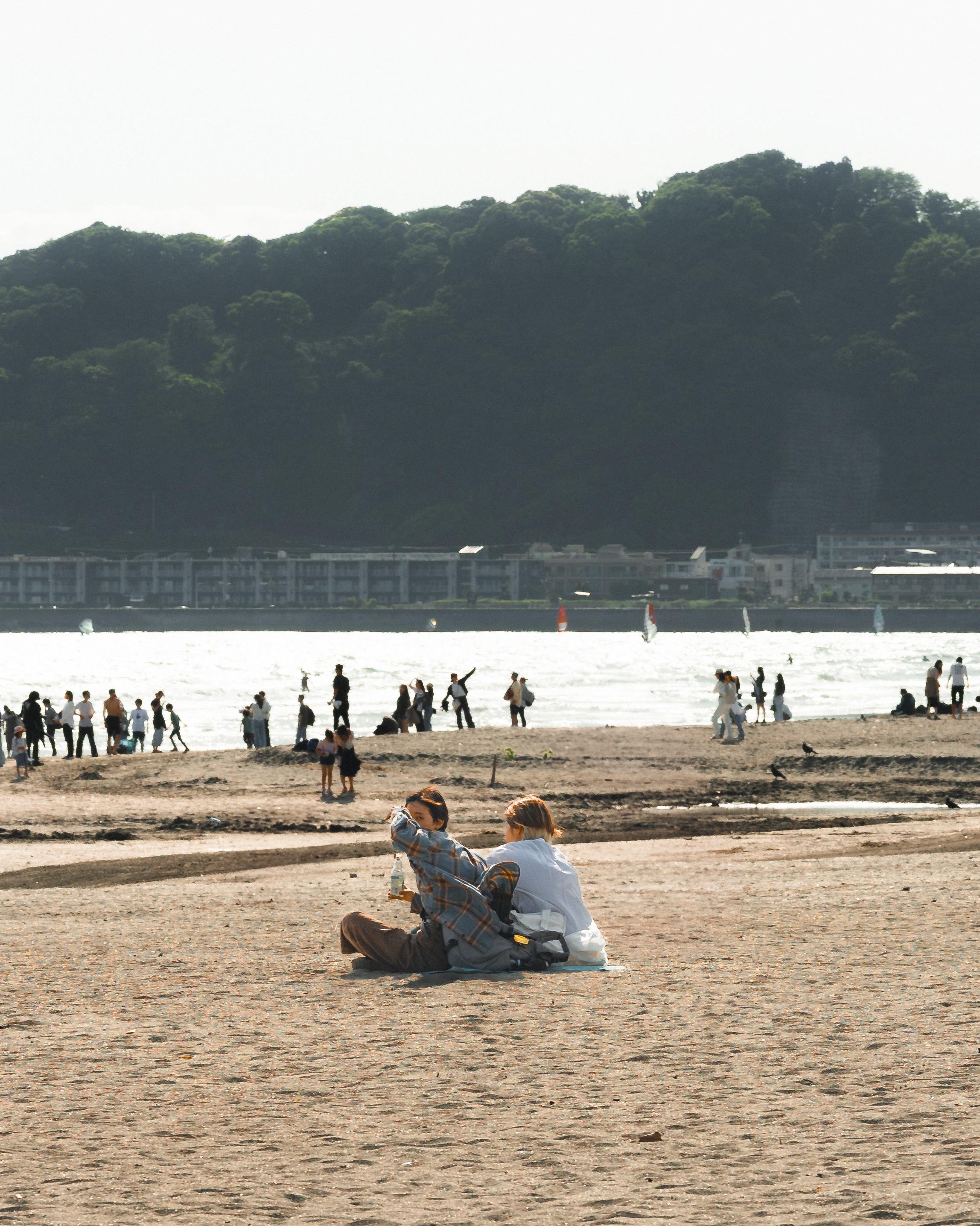 The image shows a beach scene with people relaxing and walking on the sand. In the foreground, a couple is sitting on the beach with some personal items next to them. In the background, more people can be seen near the shoreline. There are hills or a forested area across the water, and some buildings are visible along the coast. The atmosphere appears calm and sunny.