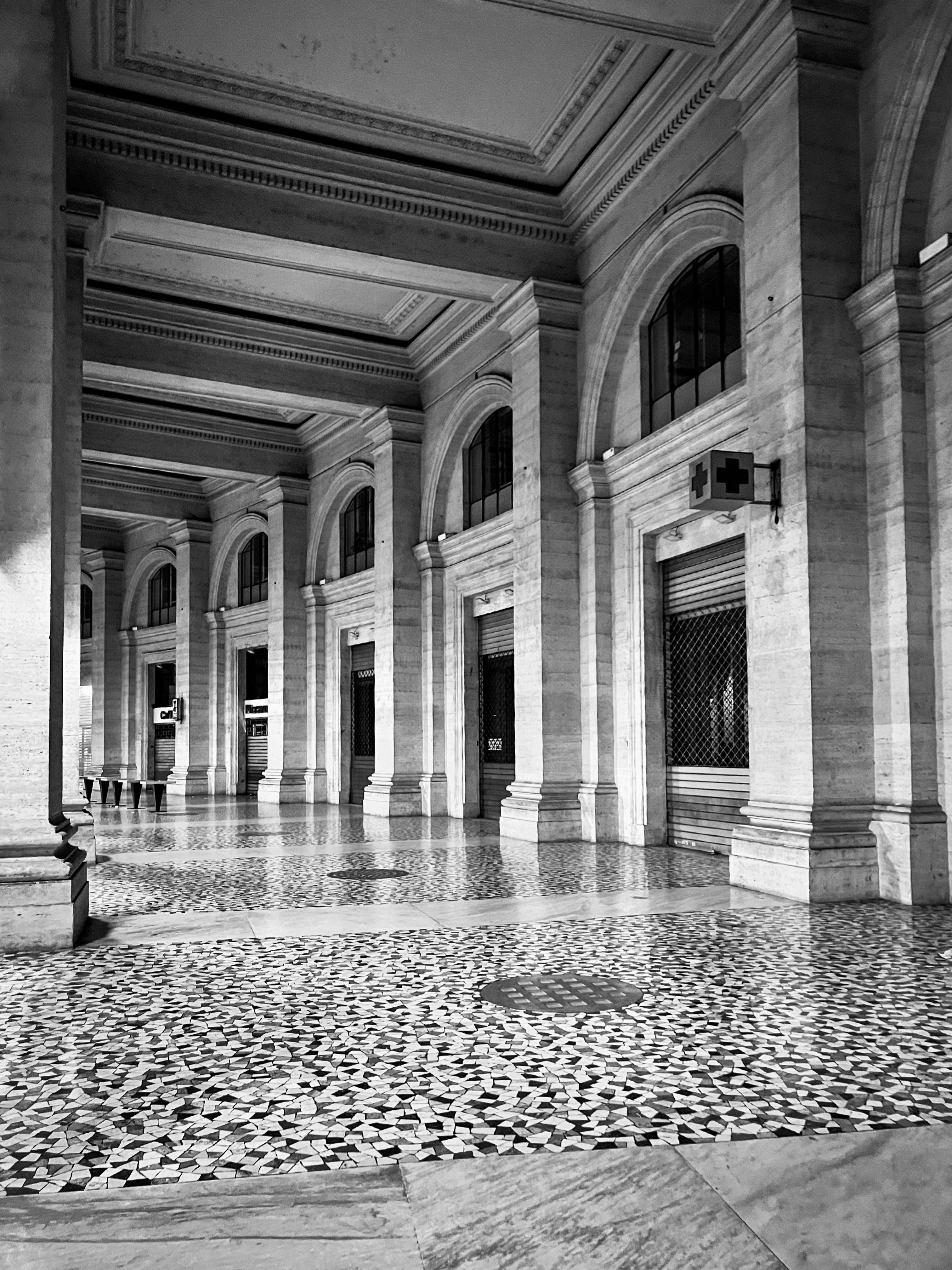 Foto in bianco e nero dell’interno di un edificio con una serie di finestre ad arco e colonne. Il pavimento ha un motivo decorativo, possibilmente fatto di piastrelle, e il soffitto presenta modanature decorative. L’architettura appare classica, con un’attenzione alla simmetria e alla grandiosità