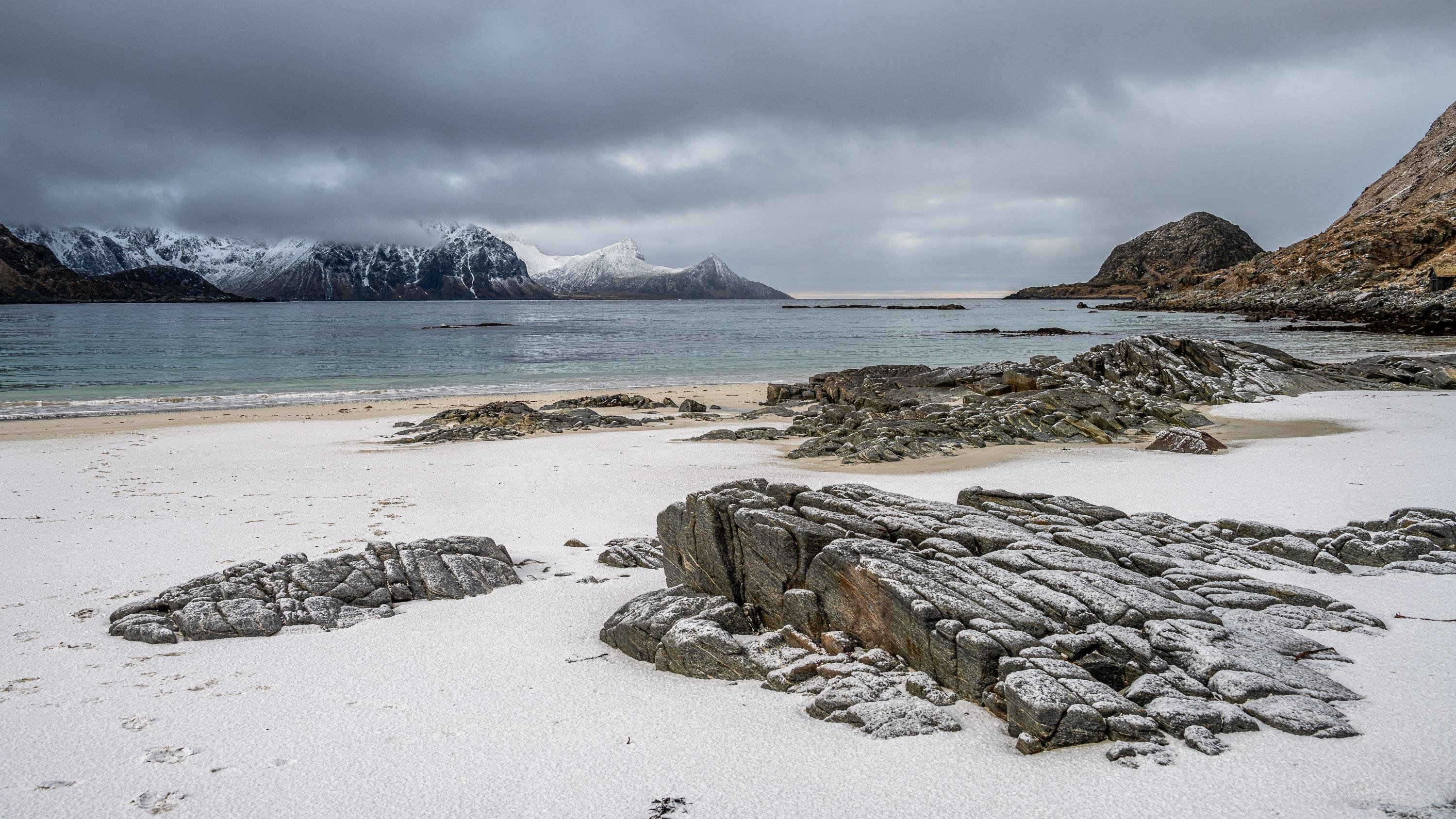 The image depicts a coastal landscape with a sandy beach, large rocks, and a mountainous background. The sky is overcast, creating a moody ambiance. The distant mountains are partially covered in snow, indicating a cold climate. The beach and rocks may also have a light dusting of snow or frost.