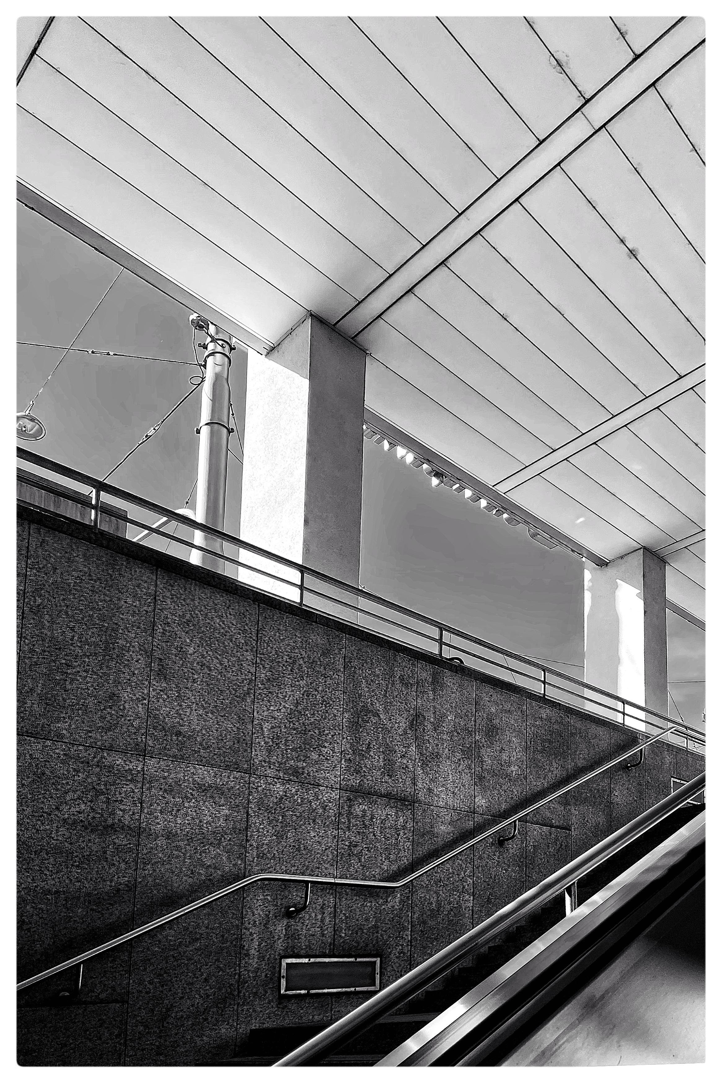 Modern architectural structure in black and white with a focus on geometric lines. Angled view of a ceiling with rectangular panels, stone wall, and metal railing. Overhead, a tall metal pole and electrical wires are visible. A staircase and escalator are partially shown, leading upward.
