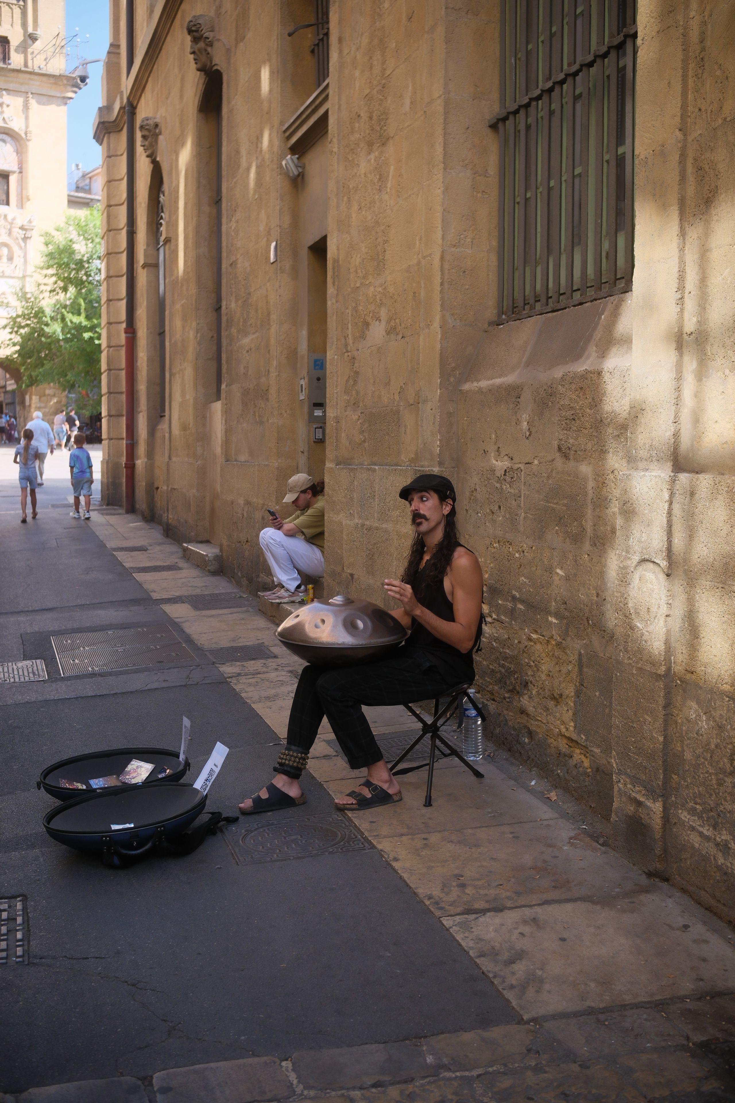 Photo verticale. Un homme assis sur un tabouret dans la rue, portant un beret noir, une moustache, un débardeur noir et un pantalon noir, jouant sur un hang