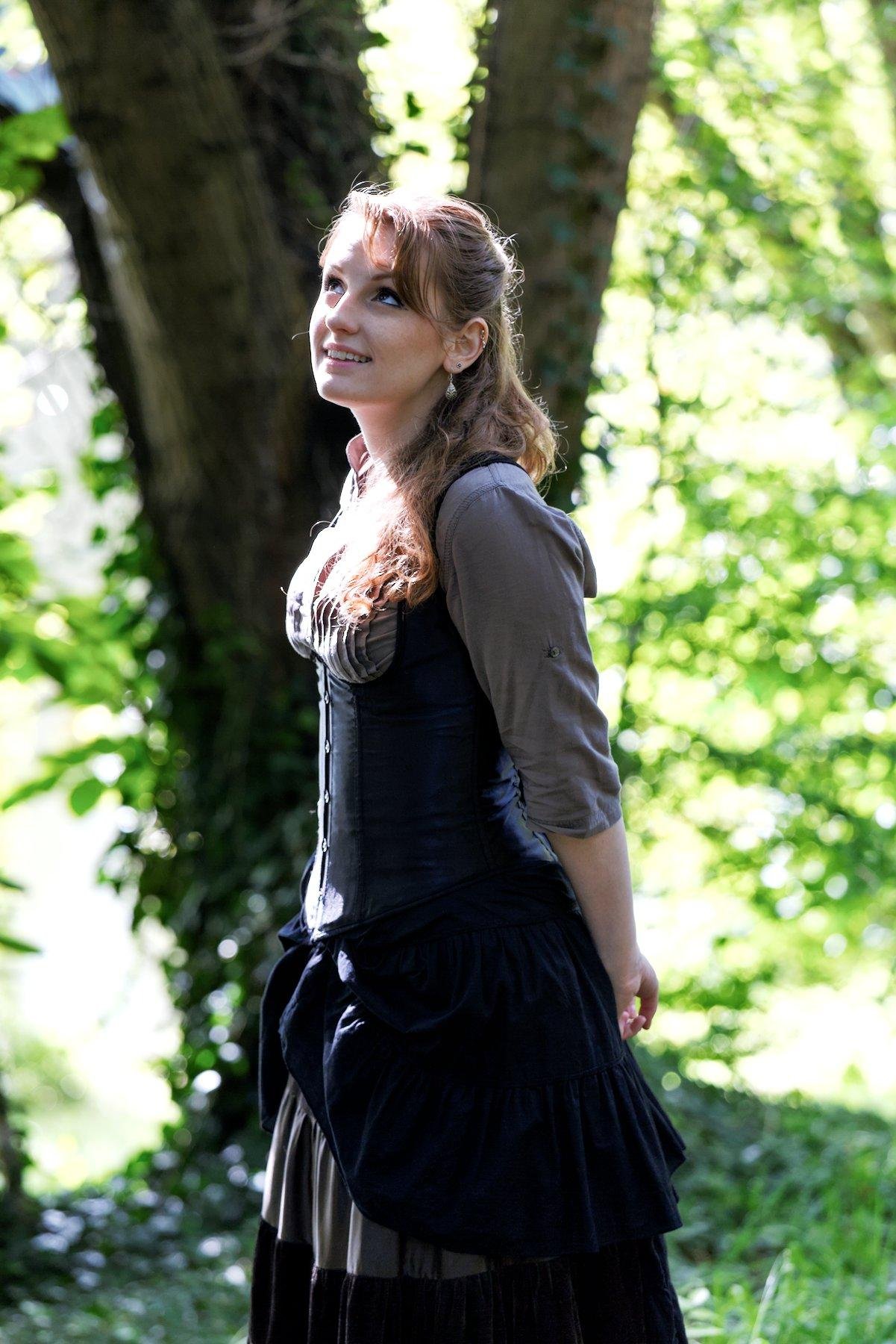 Photo portrait, montrant une jeune femme rousse au cheveux long élégamment coiffé, portant un chemisier marron et par dessus, un corset noir, une longue jupe marron foncé. Elle se tiens debout, les mains derrière le dos, regardand en l’air en souriant. Le fond est composé d’un gros arbres et de feuillages verts