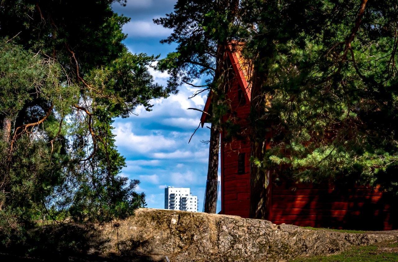 Red wooden building partially obscured by tall pine trees in the foreground, with a distant view of a modern white high-rise building under a cloudy blue sky. A rugged stone wall or path runs across the lower part of the image.