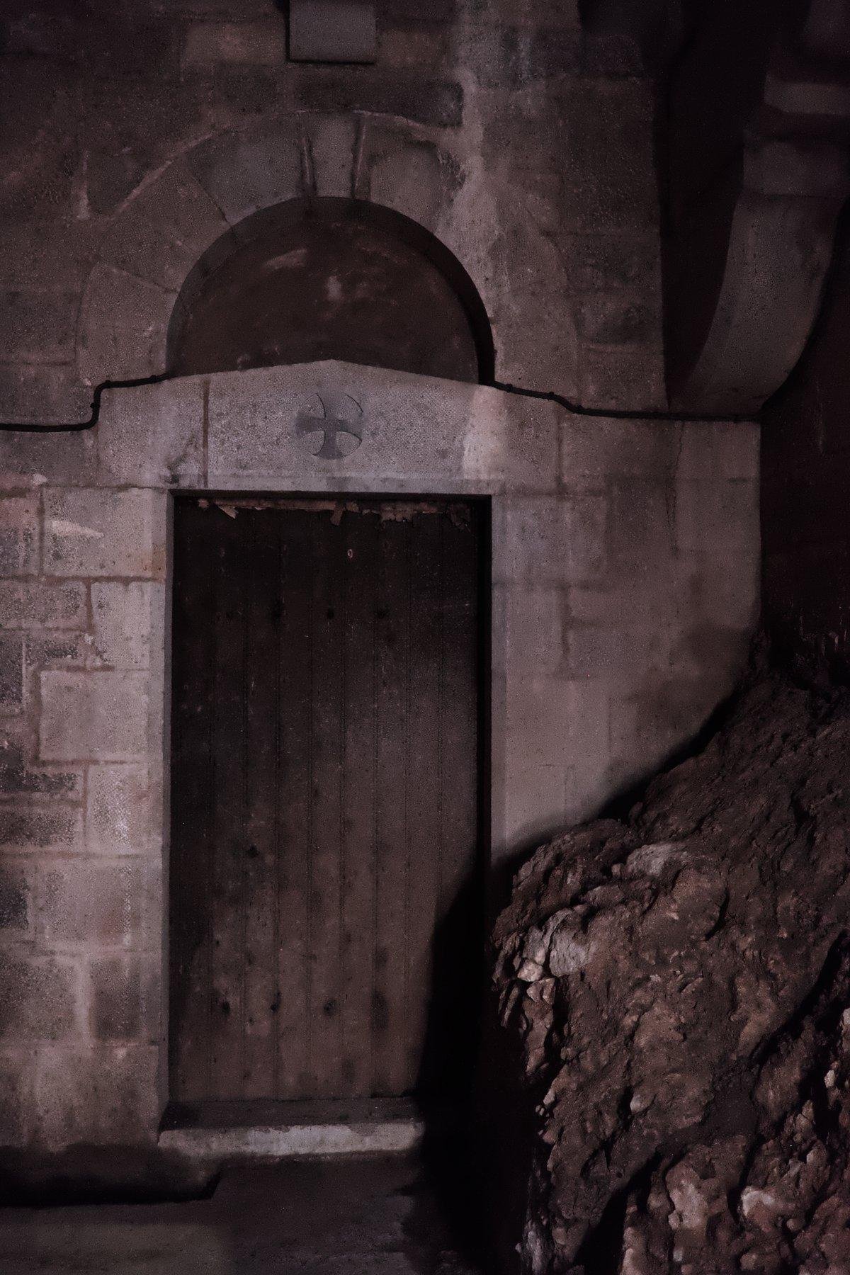 Photo verticale. Une porte en bois avec une croix gravée au dessus dans le mur de pierre. Elle est un peu cachée par de la roche