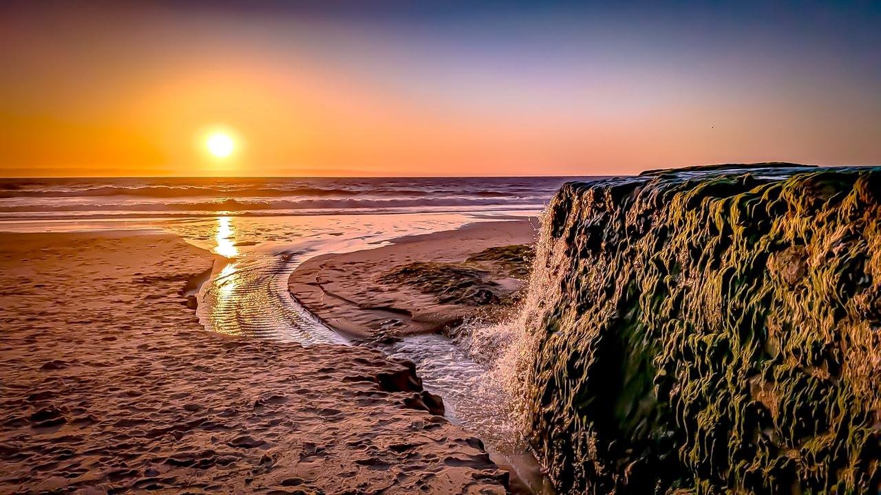 Sunset over the ocean with a glowing sun near the horizon, casting reflections on the water. A small stream flows from the foreground into the ocean, bordered by sandy shores. On the right, a rocky outcrop is covered in green moss, with water gently cascading down its surface. The sky transitions from deep blue to warm orange near the horizon.