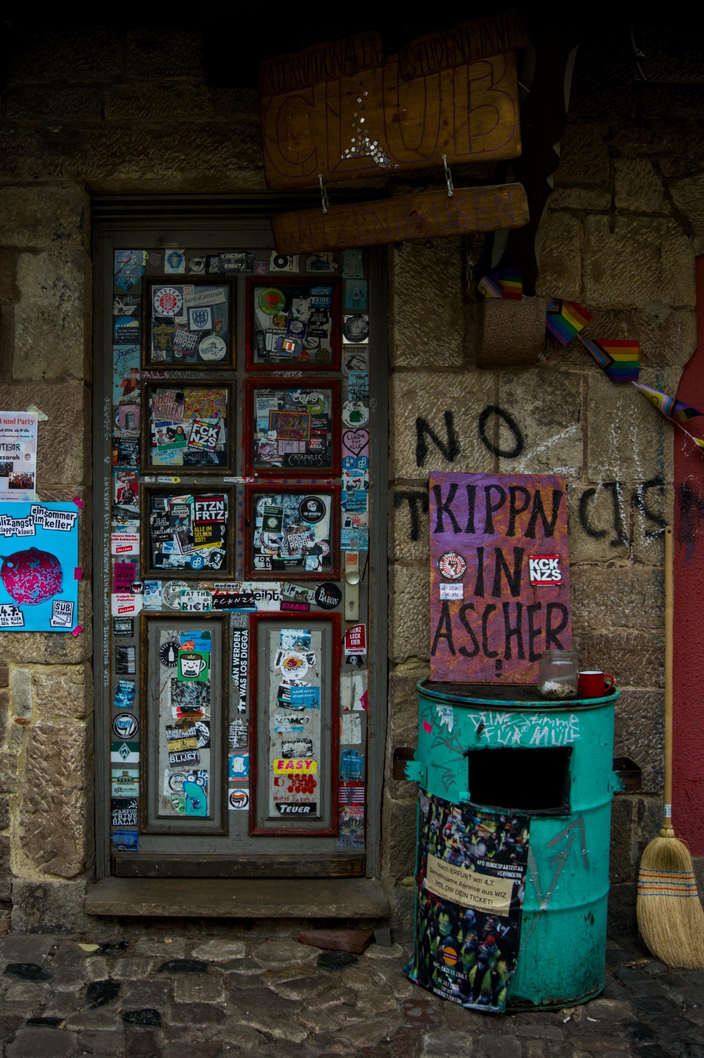 Stone-walled entrance with a door covered in stickers, topped by a hanging wooden sign. To the right, graffiti and a handwritten sign reading “KIPPN IN ASCHER” resting on a green, graffiti-marked trash can and a broom leaning against the wall.