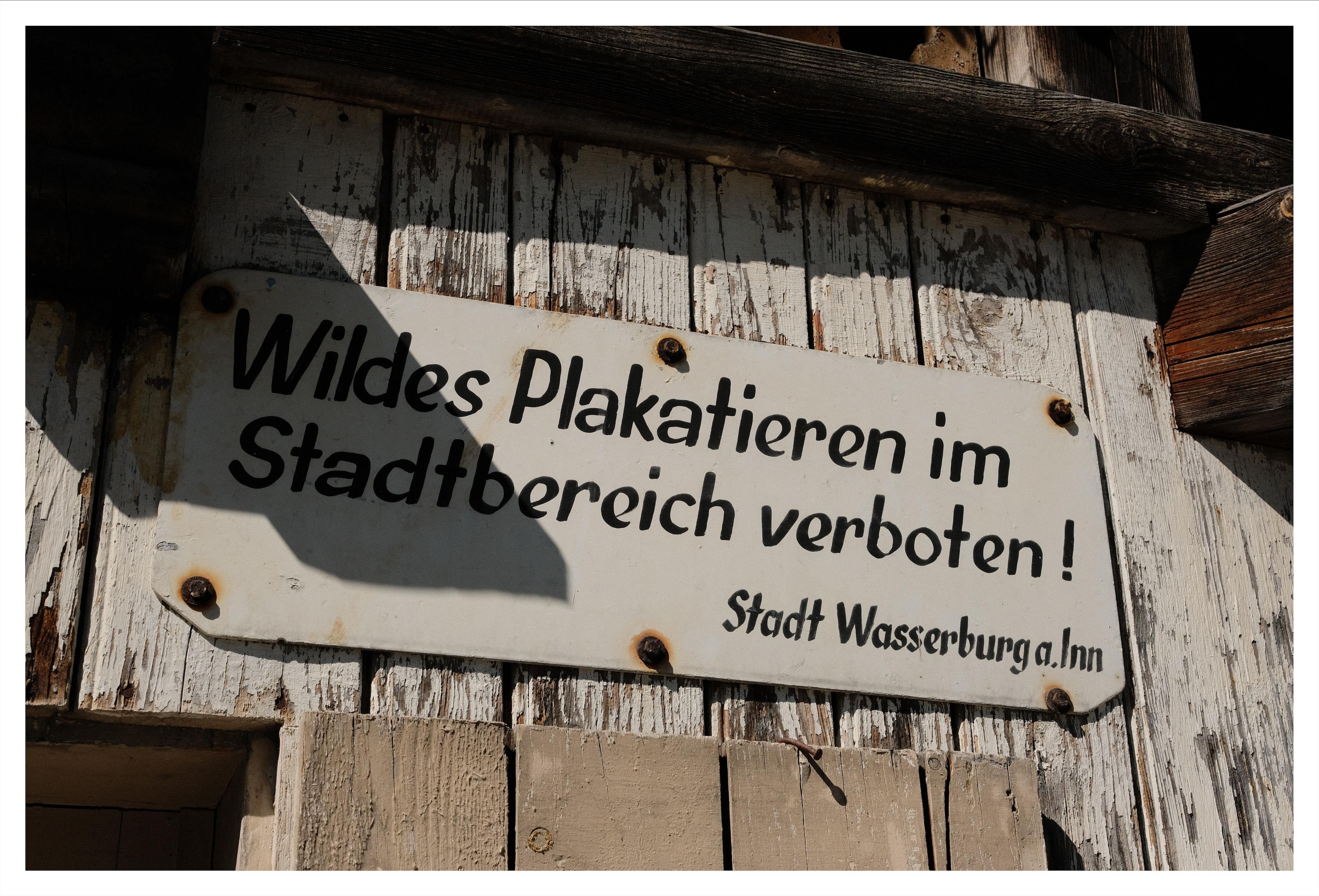 White metal sign with black German text “Wildes Plakatieren im Stadtbereich verboten!” and smaller line “Stadt Wasserburg a. Inn,” screwed to a weathered wooden wall with peeling paint and shadows across the sign.