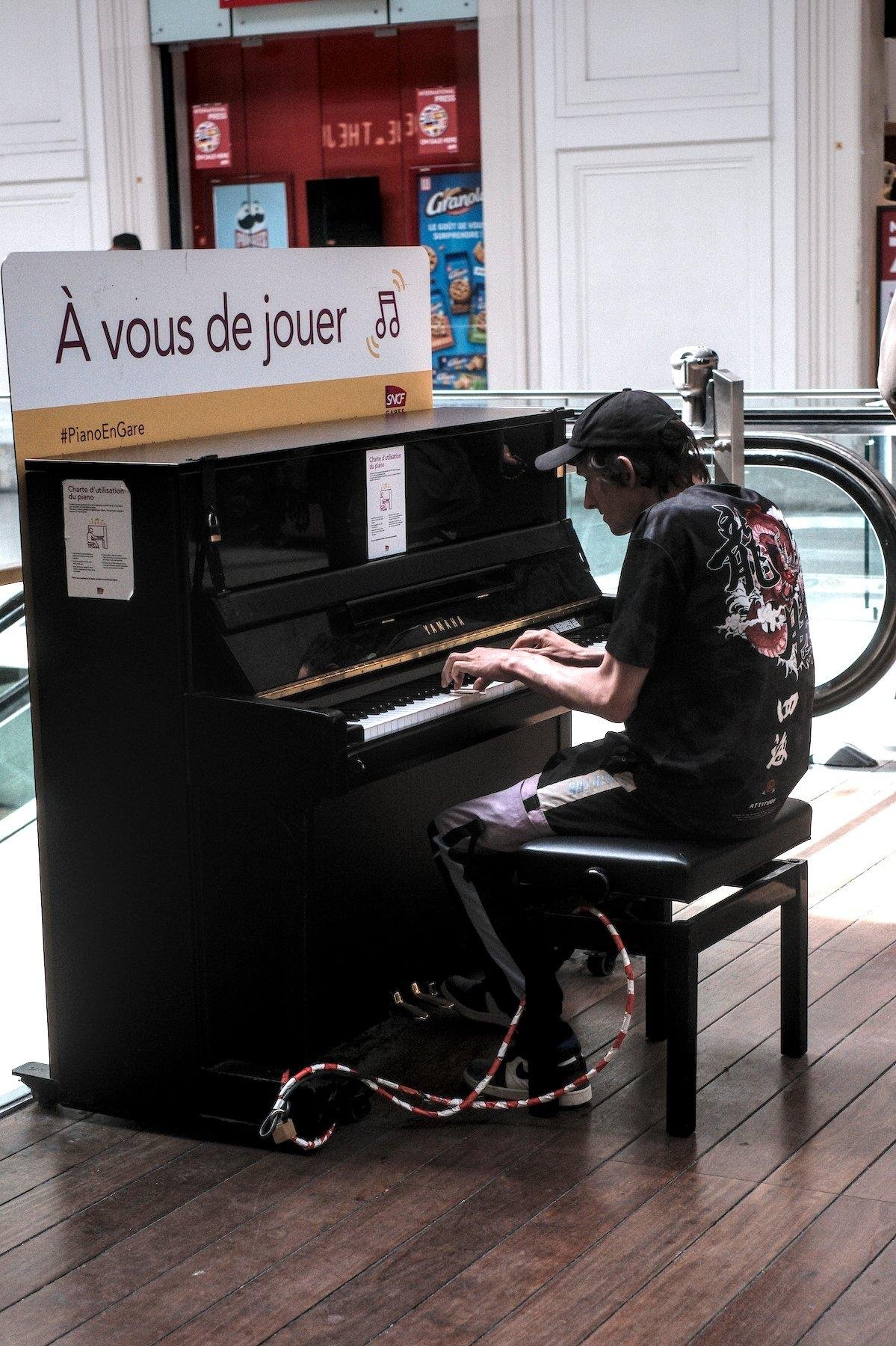 Photo verticale. Dans un lieu publique, un piano droit est à disposition, avec une pancarte au dessus marqué «&nbsp;À vous de jouer&nbsp;». Un jeune homme brun, casquette noire, t-shirt noir, 3/4 de dos, est entrain de jouer