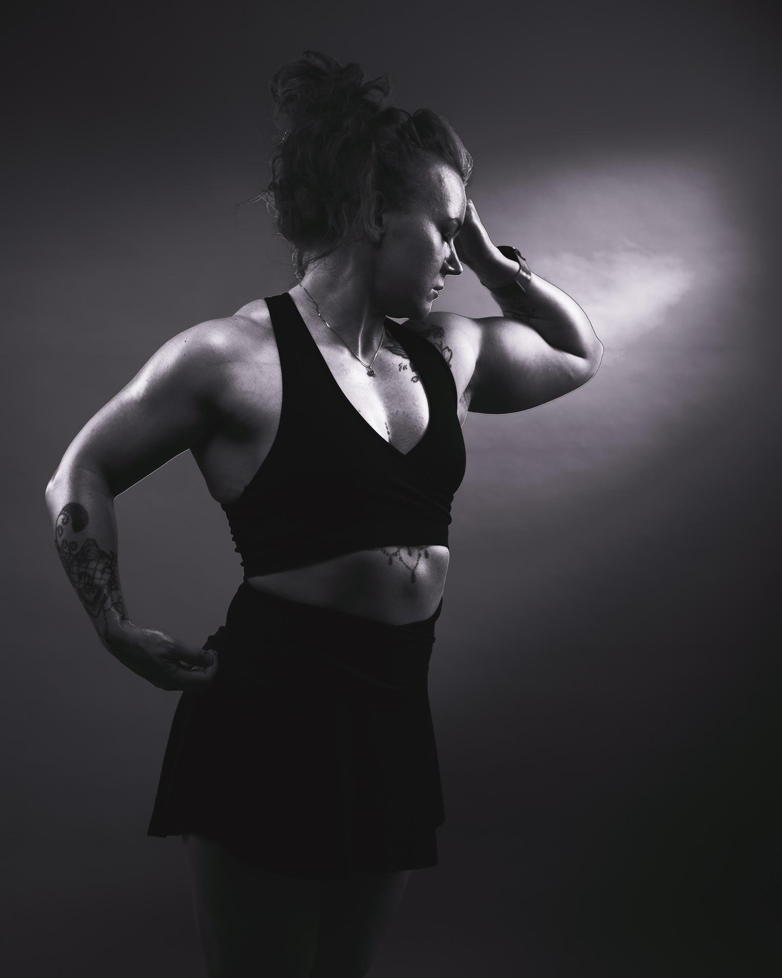 The image shows a black-and-white photograph of a person posing in an athletic stance. They are wearing a sleeveless top and a skirt. The person is holding one hand on their hip and the other on their head, showcasing their muscular arms. The lighting creates a dramatic effect by highlighting their physique.