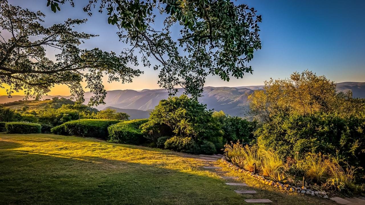 The image features a scenic landscape with a well-maintained garden in the foreground. There's a grassy lawn with a stone path leading through it. The area is surrounded by lush greenery, including various shrubs and trees. In the background, there are hills or mountains under a clear blue sky. The lighting suggests it might be early morning or late afternoon, creating a serene and picturesque atmosphere.