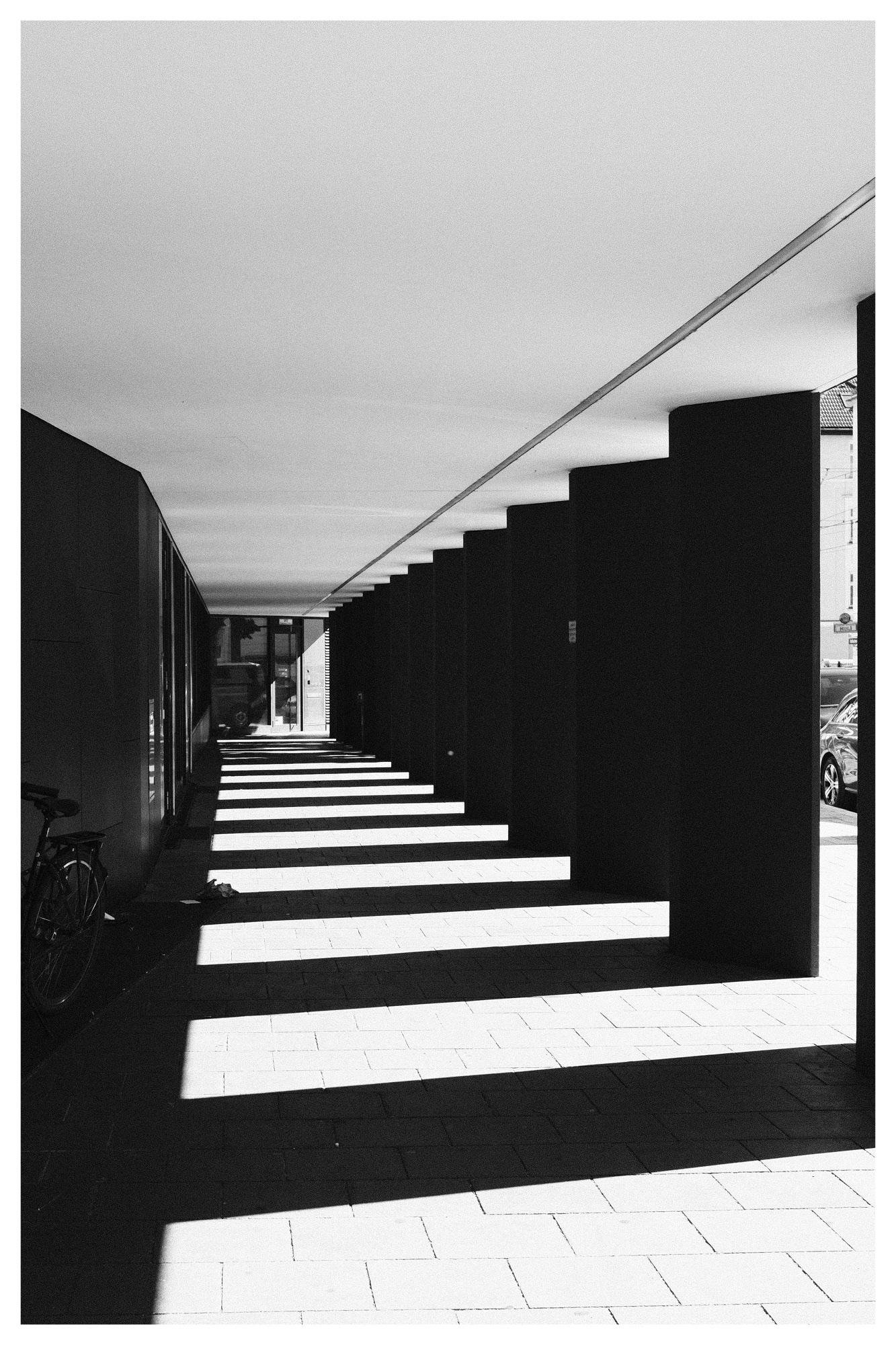 Black-and-white view down a covered walkway with a row of rectangular columns on the right casting long striped shadows across the tiled floor; dark wall on the left with a bicycle partially visible, and a street with parked cars at the far right edge near the exit.