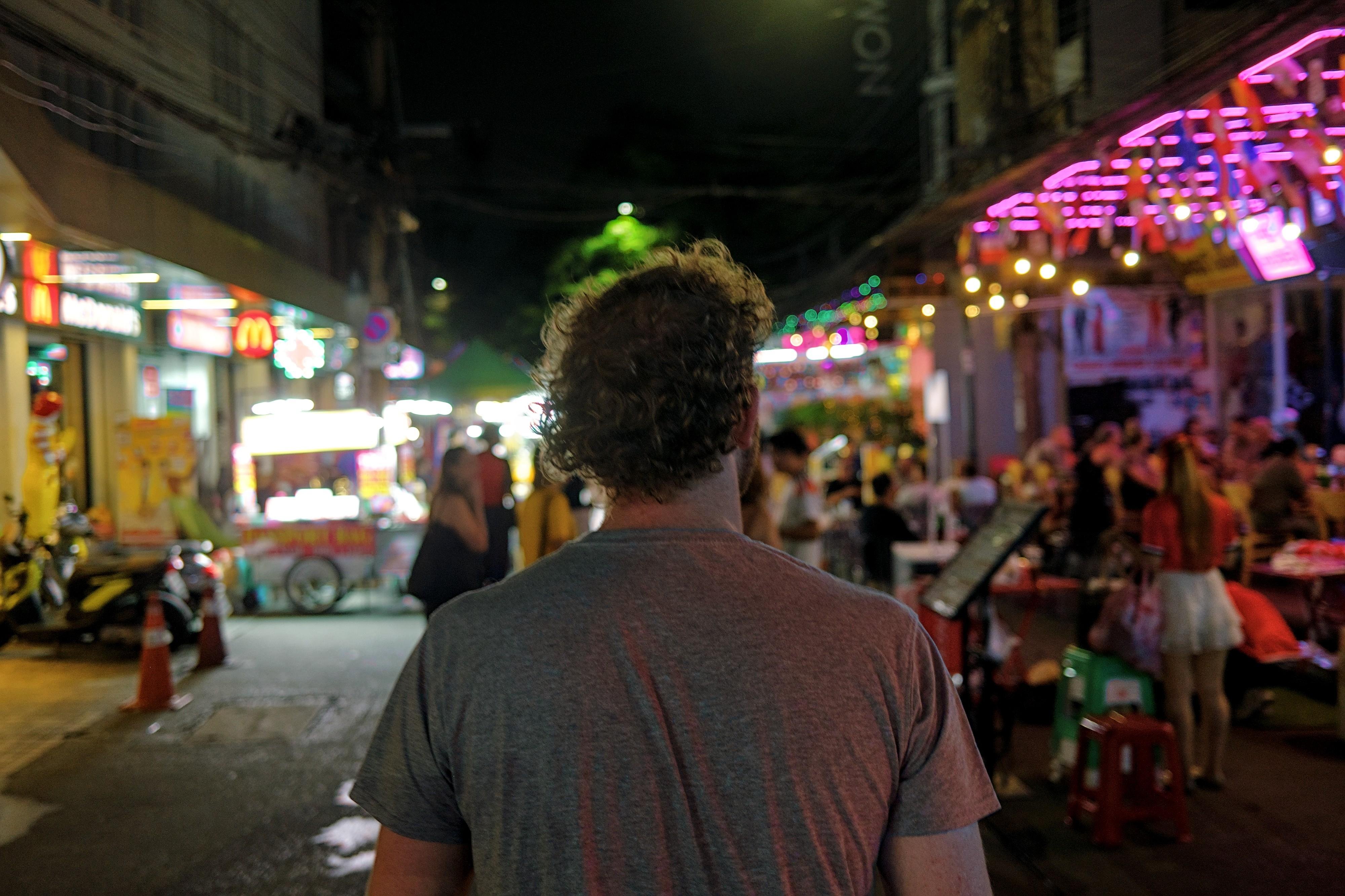 Rear view of a person with curly hair in a gray T-shirt walking down a busy night street lined with neon-lit bars and restaurants, with people seated at outdoor tables and bright storefront signs on both sides.