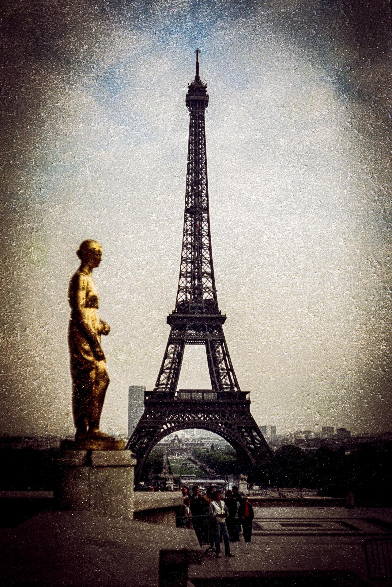 A textured image featuring the Eiffel Tower centered in the background with a golden statue on the left in the foreground. Several people walk in front of the tower, and the sky is overcast, lending a vintage or grainy feel to the scene.