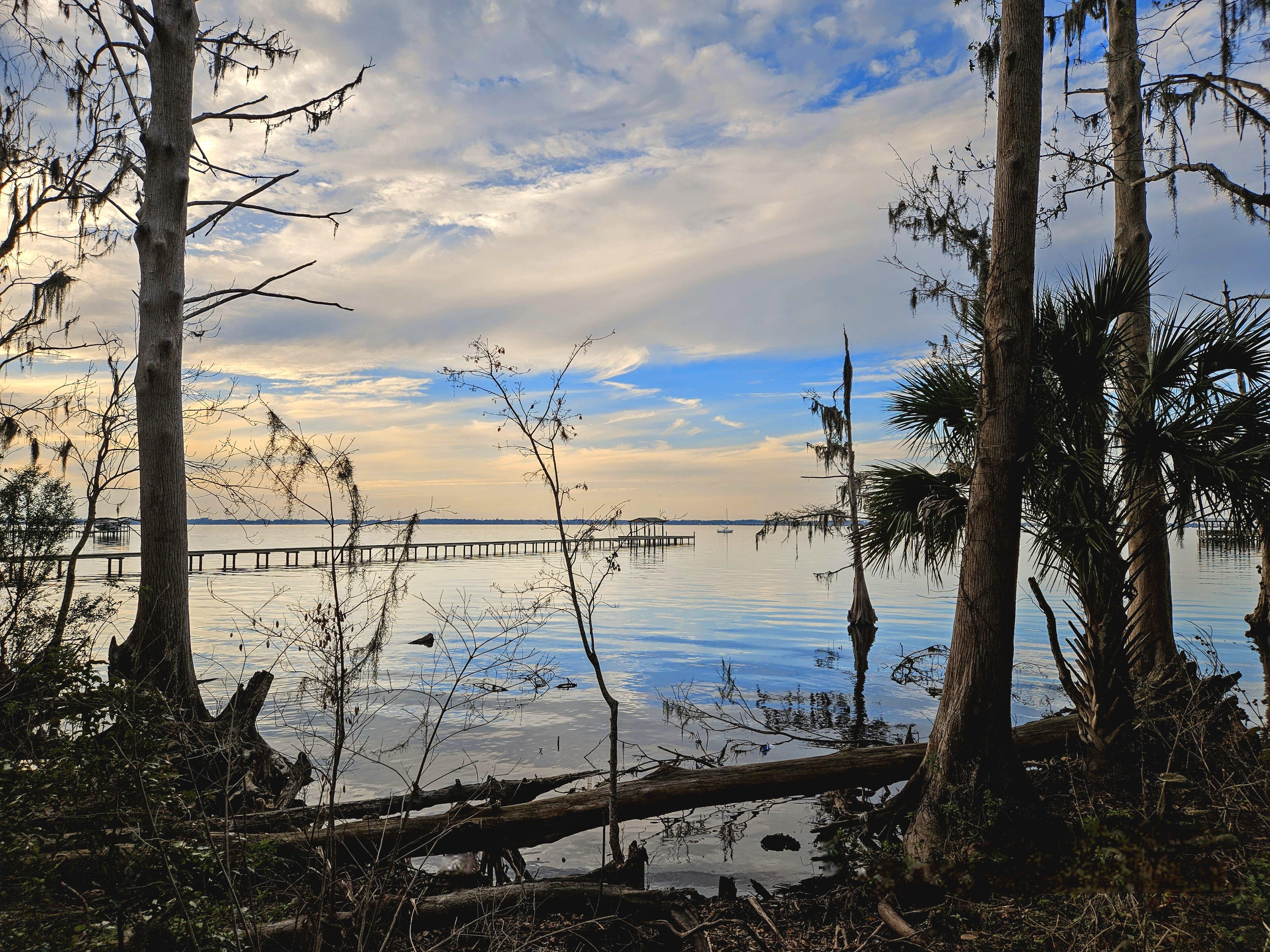 Trees and foliage frame a tranquil waterfront scene at sunset, with a calm body of water reflecting the sky. A wooden pier extends into the water, while clouds display soft hues of blue, white, and orange in the background.
