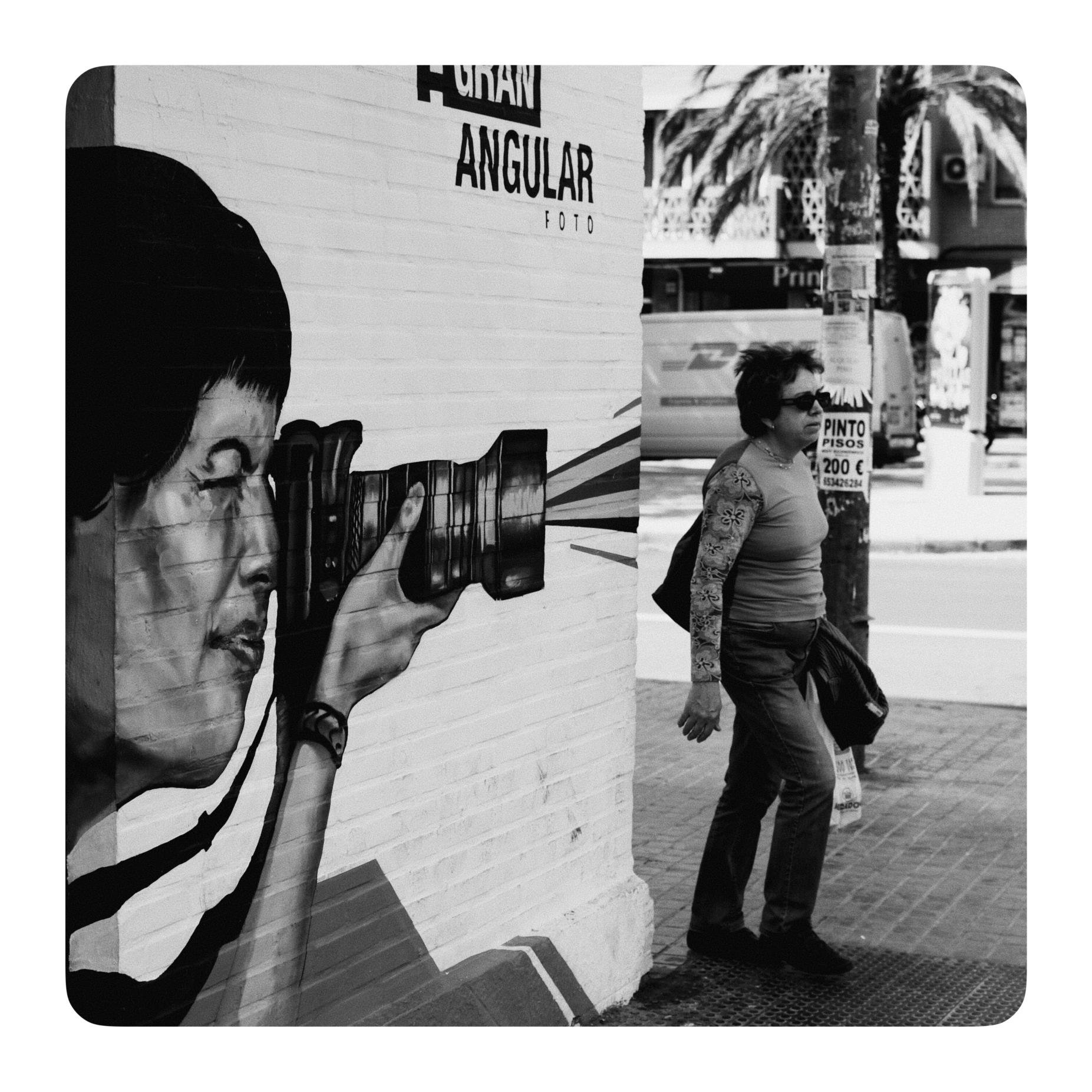 Graffiti von einer Person die eine Kamera hält und durch den Sucher schaut. Daneben geht eine Frau vorbei. Es sieht aus als würd diese vom Graffiti fotografiert werden.