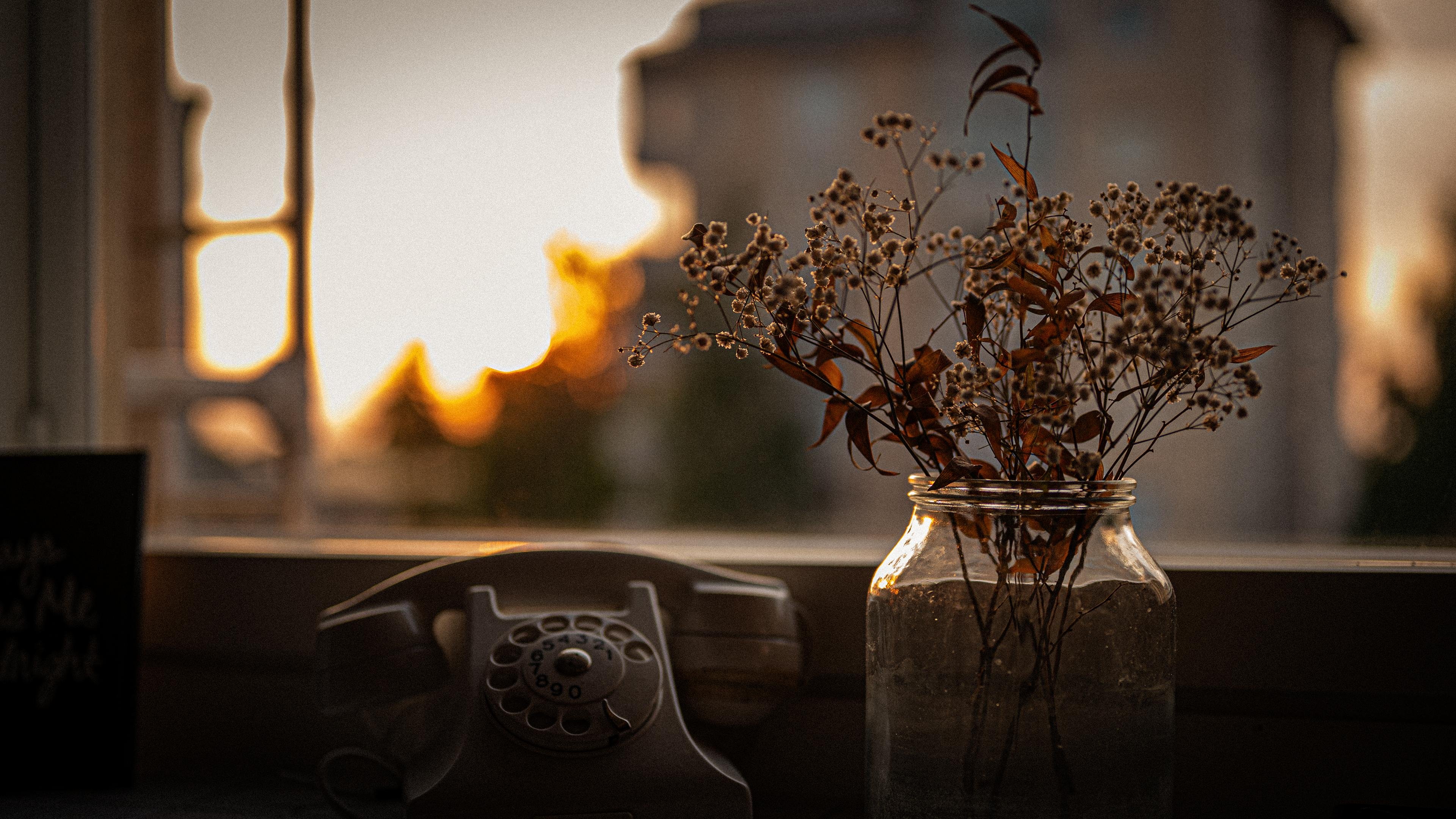 Rotary phone and a jar with dried flowers on a windowsill, with a warm, blurred sunset view outside. A building is visible in the distance, softened by the glowing light. The overall tone is autumnal and serene.