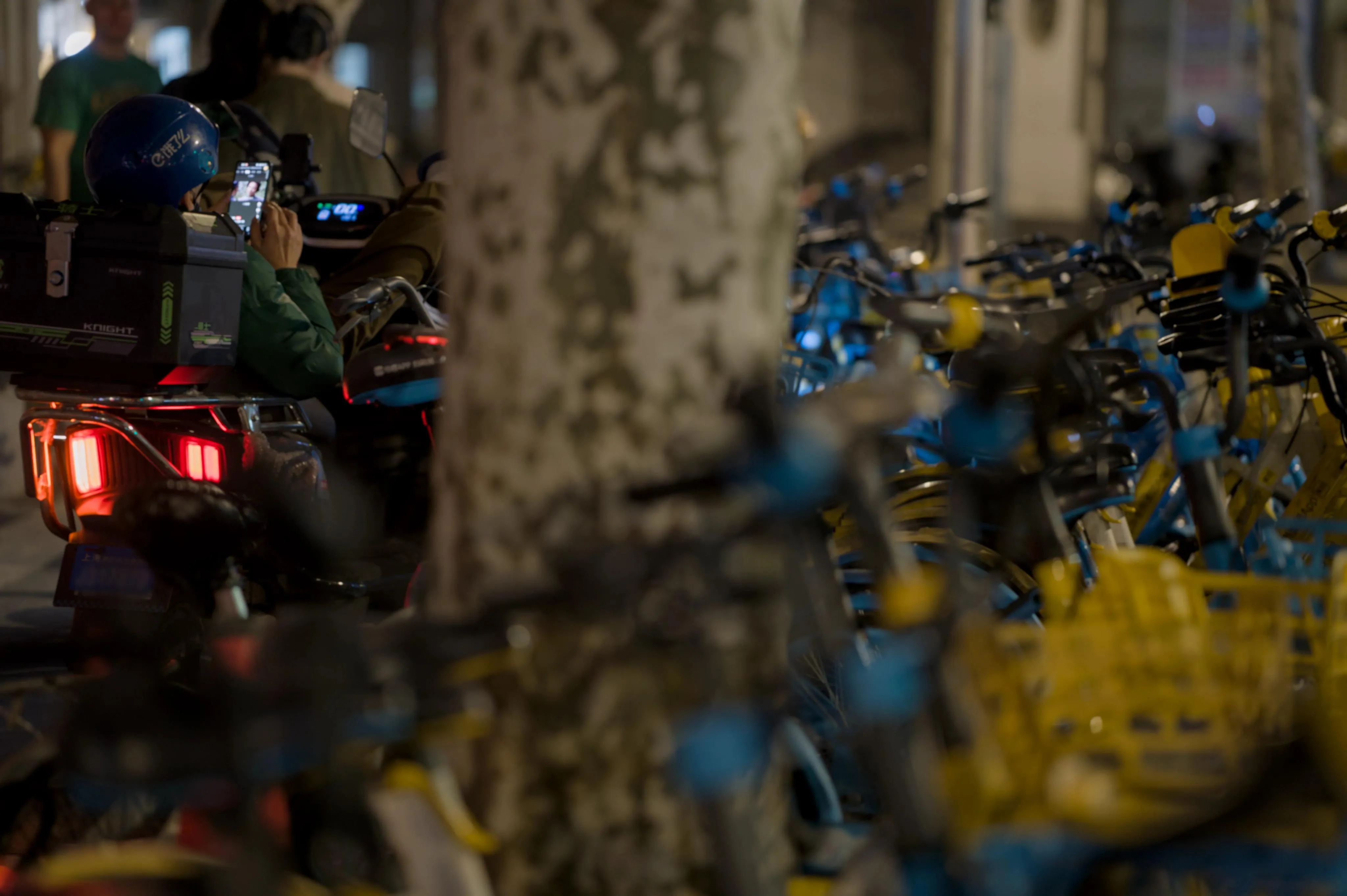 Nighttime photograph of public-use bicycles stationed around a tree trunk. The bicycles are blue and yellow in color, with yellow baskets on their front. On the other side of the tree trunk, a delivery driver waiting on his scooter and using his smartphone, with his feet on the scooter's dashboard. 