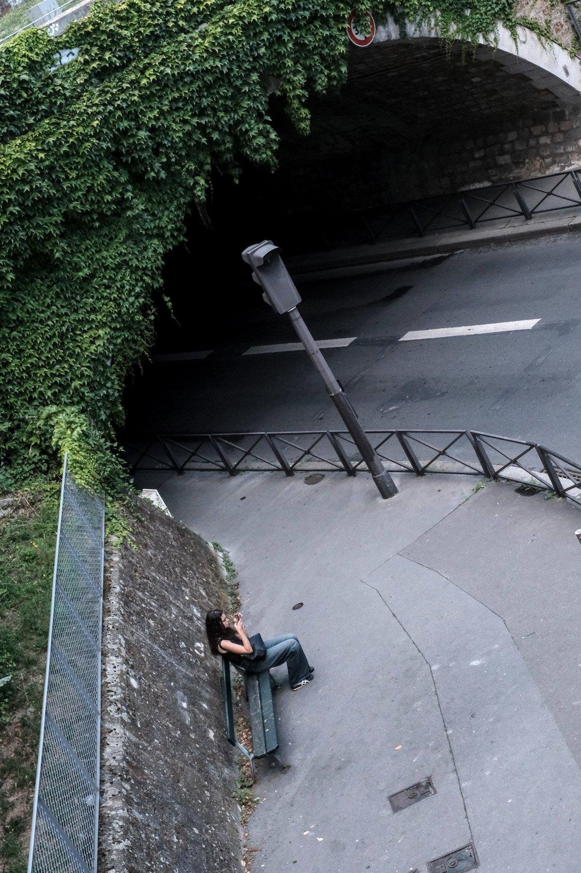 Photo verticale. Plongée sur une rue, un banc vert contre un mur pas très haut se finissant sur un grillage protégeant une pelouse à gauche. En haut de l’image l’entrée d’un tunnel routier avec le mur extérieur couver de lierre bien vert. Sur le banc, une jeune femme assise en jean et débardeur noir, cheveux noirs long et lâchés, consultant son téléphone l’air agacée